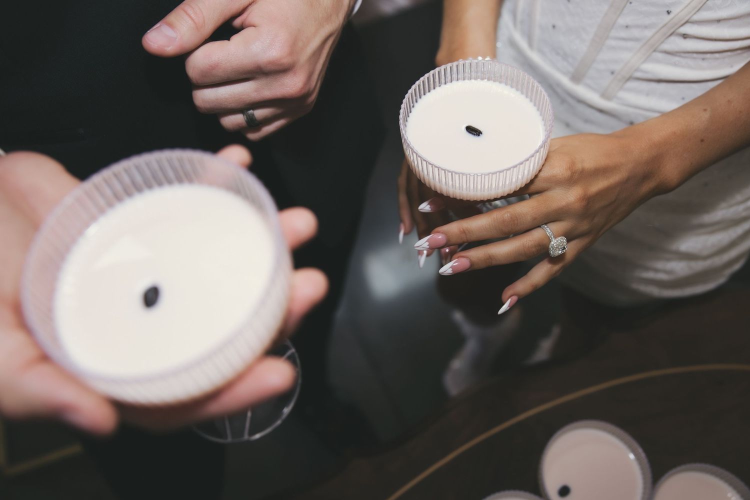 Hands holding white glass cocktail beverages with dark seeds floating on top of the creamy foam.