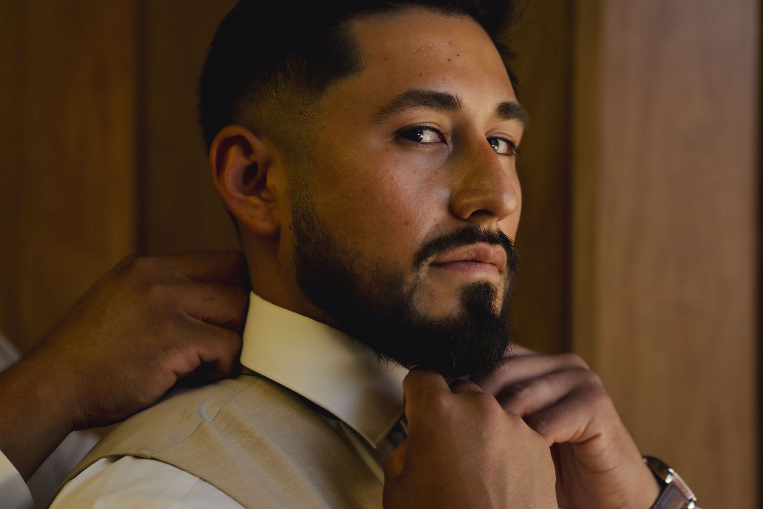 Someone adjusts a bowtie while wearing a cream-colored formal suit jacket in warm lighting.