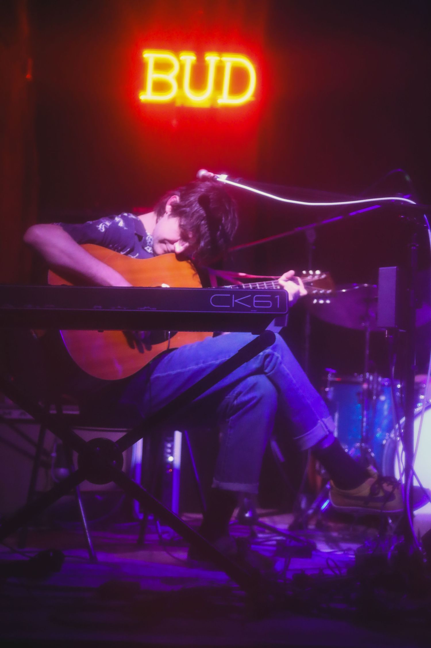 Musical performance in dark venue with red neon Bud sign glowing and musicians playing instruments under purple lighting.
