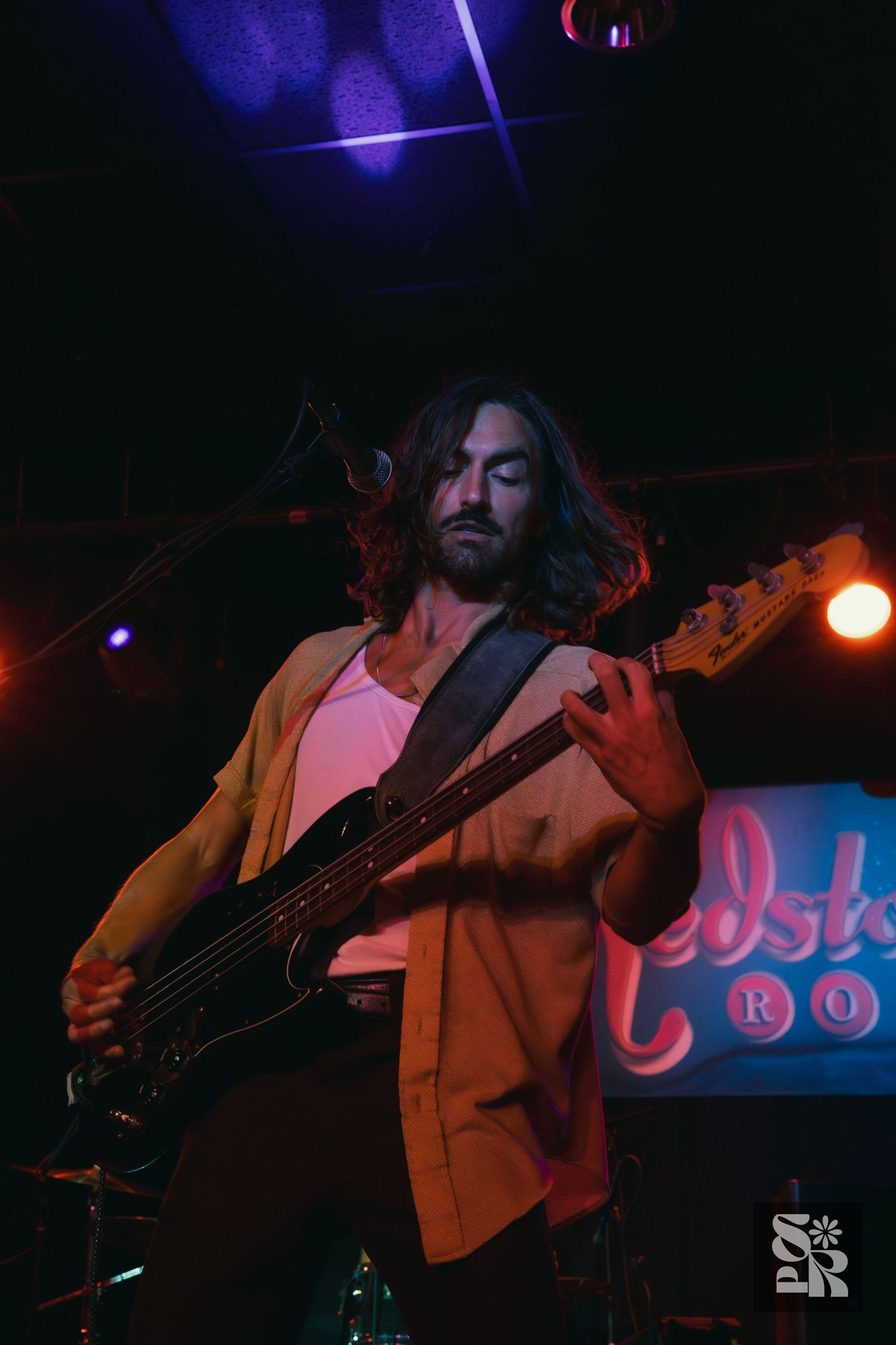 Bass player performs on stage at a nightclub with purple and orange lighting while wearing a loose orange shirt.