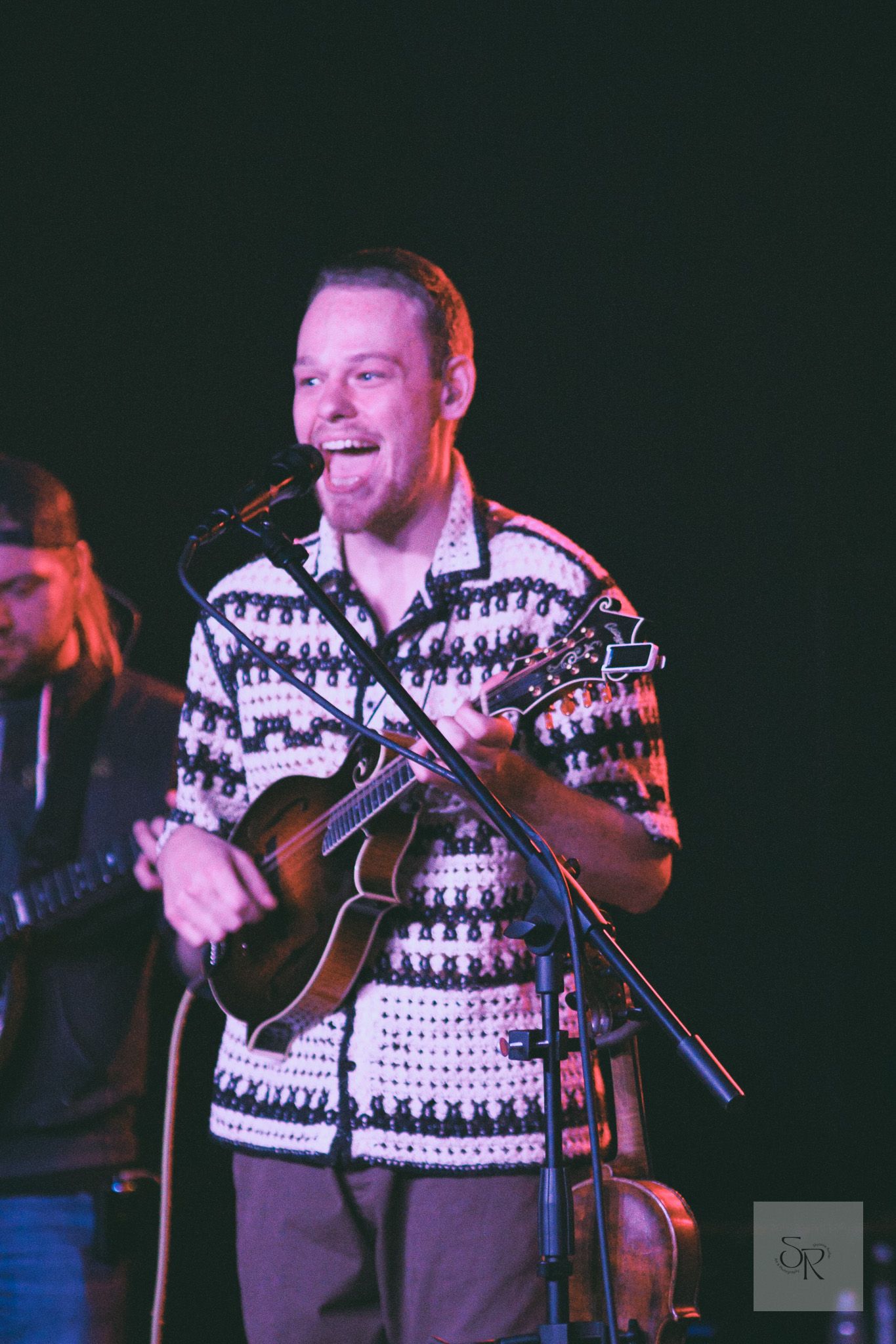 A performer in a patterned winter sweater plays acoustic guitar on stage under colorful lighting during a show.