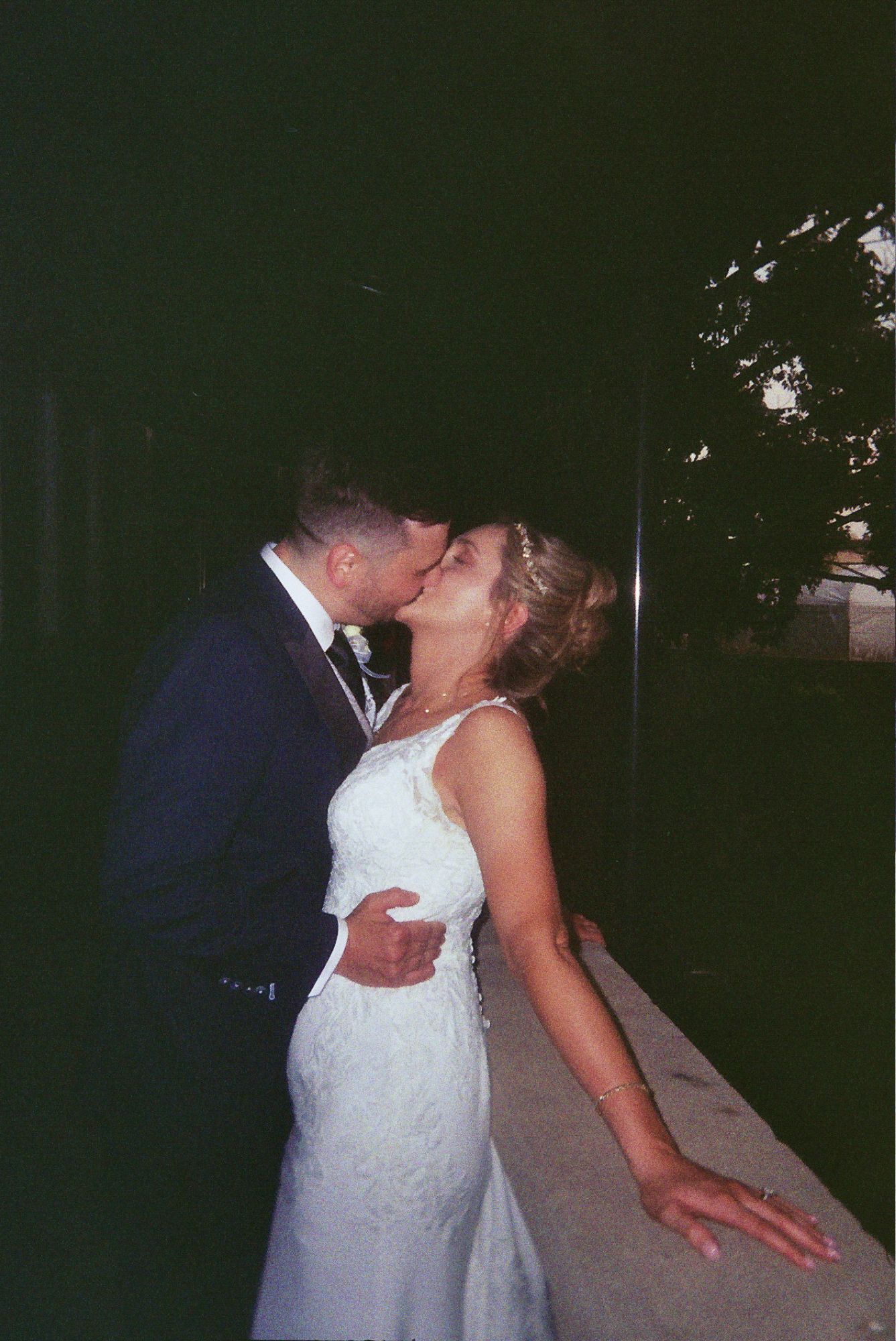 Newly married couple sharing a romantic kiss while leaning against a ledge at their outdoor wedding reception.