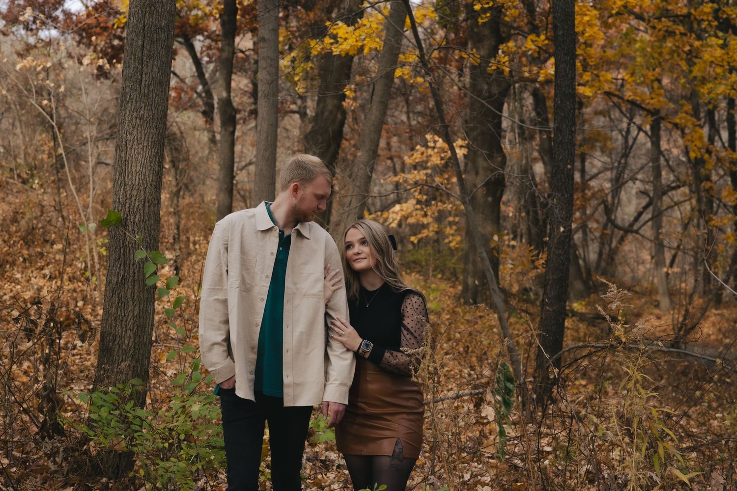 A couple embraces in a romantic autumn forest setting surrounded by golden leaves and tall trees.