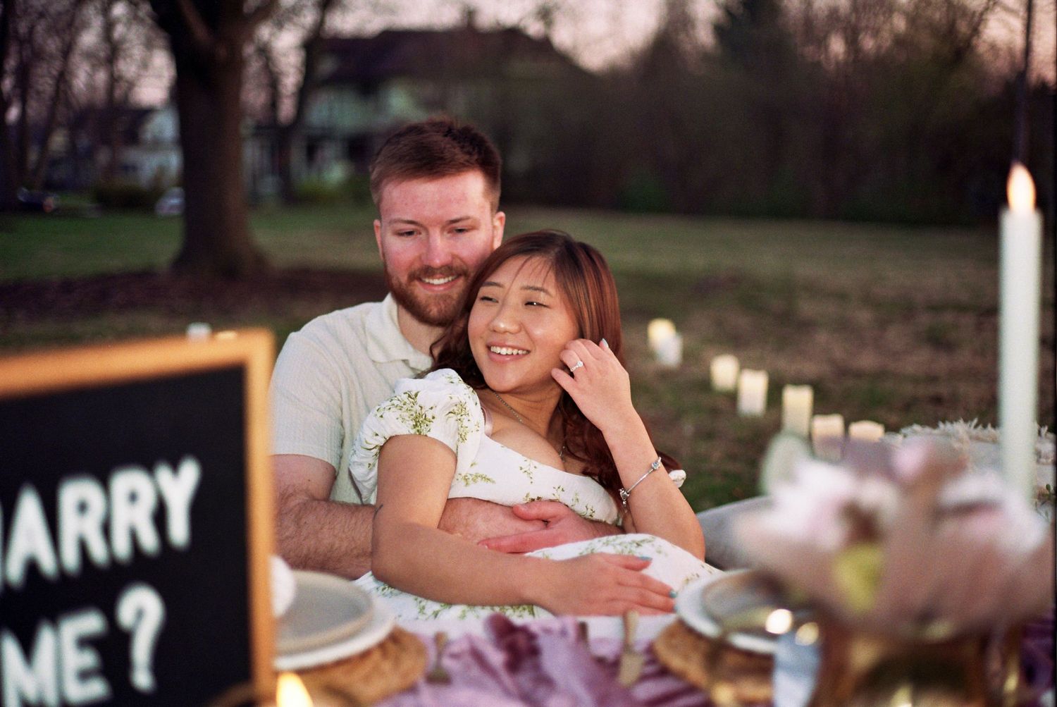 A couple shares a happy moment during an outdoor proposal setup with a 'Marry Me' sign and romantic decorations.