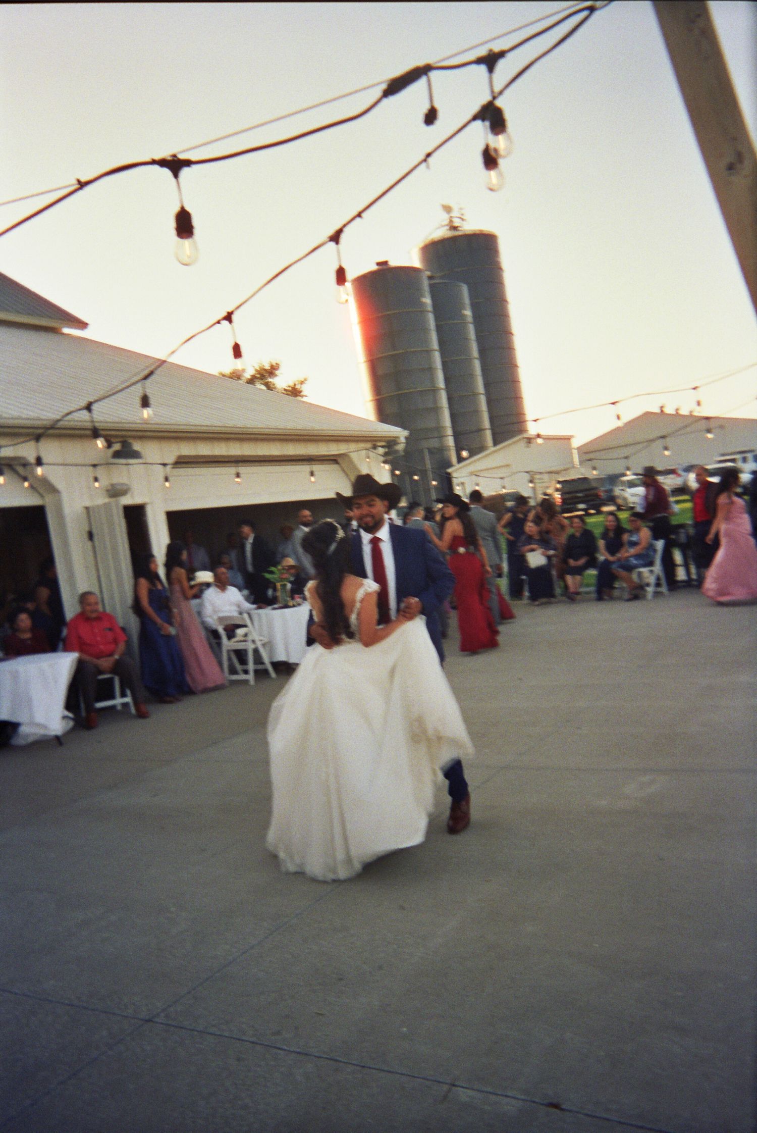 Wedding celebration on an outdoor patio with string lights and farm silos visible in the background at sunset.