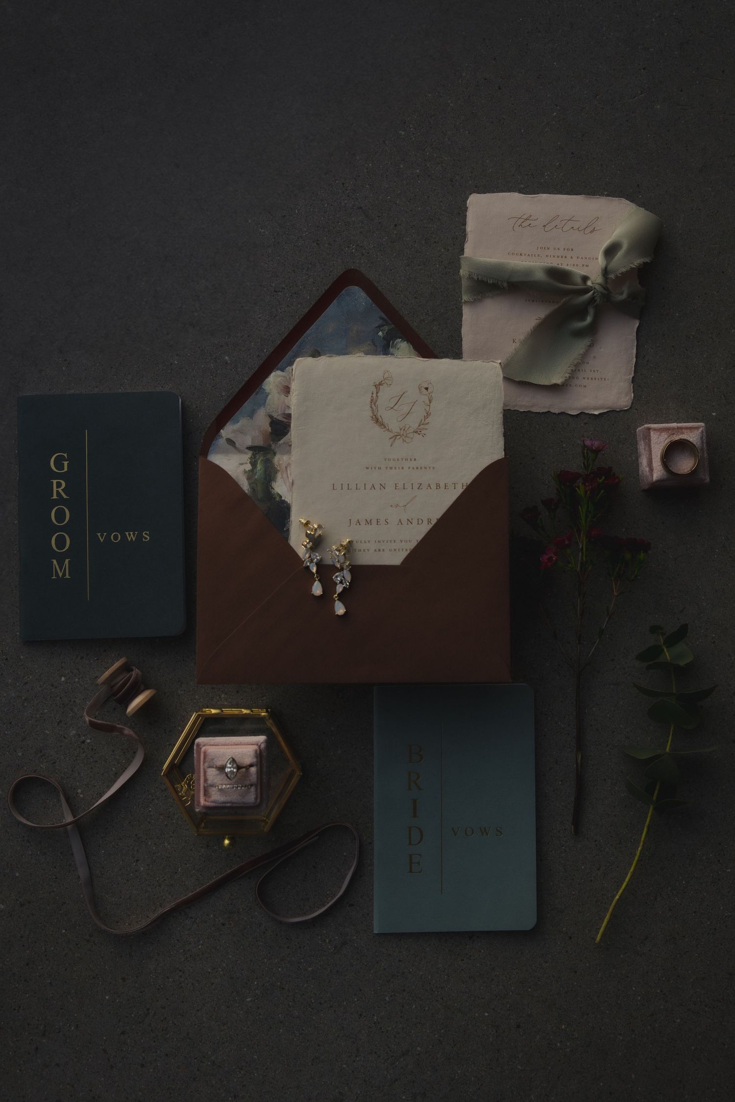 Wedding invitation suite with brown envelope, blue books, ribbon, and foliage on dark background.