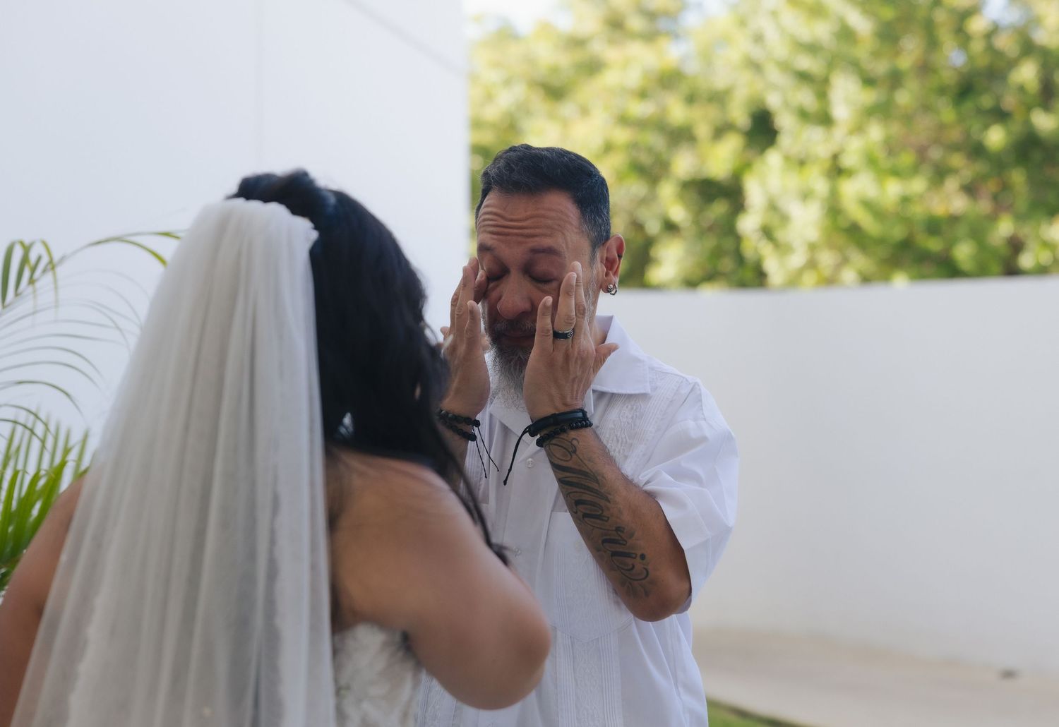 Emotional groom wiping tears during wedding first look with bride in white dress and veil outdoors.
