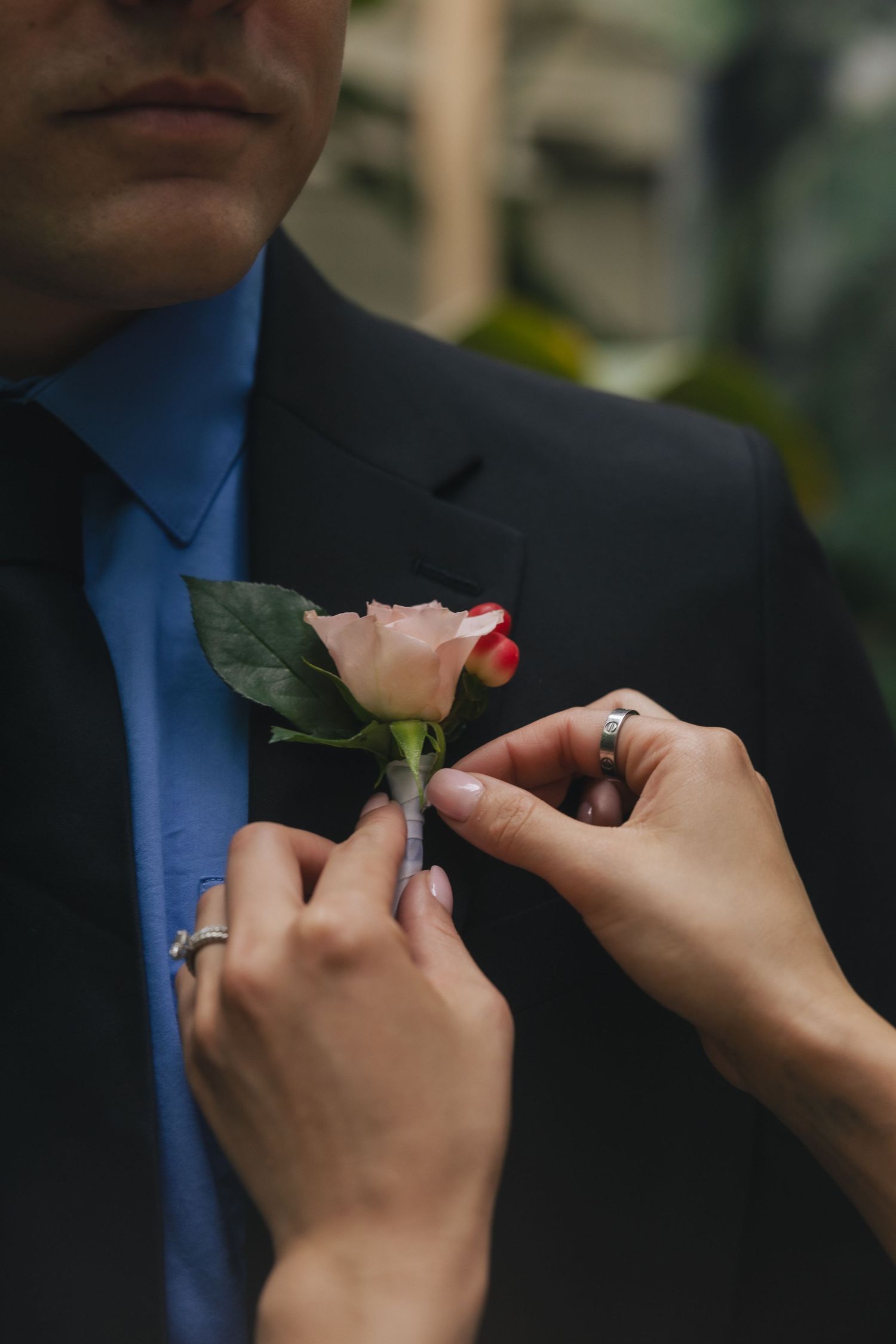 Woman with red nails pins a pink rose boutonniere on a man in a black suit and blue tie.