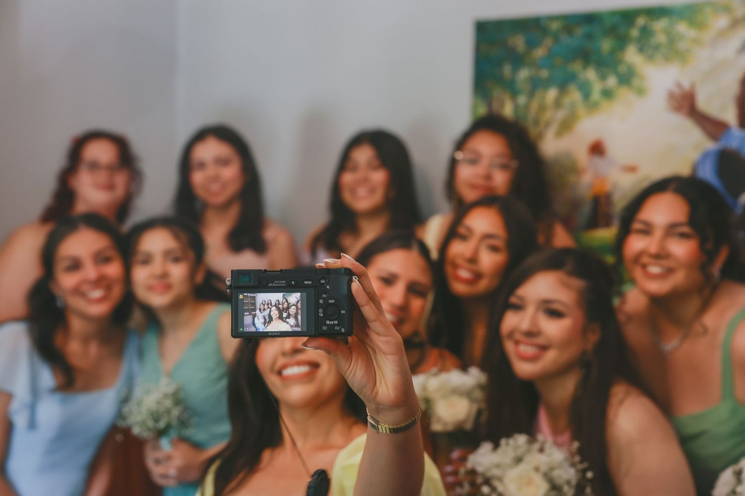 A group poses together while one member takes a selfie with a digital camera in front.
