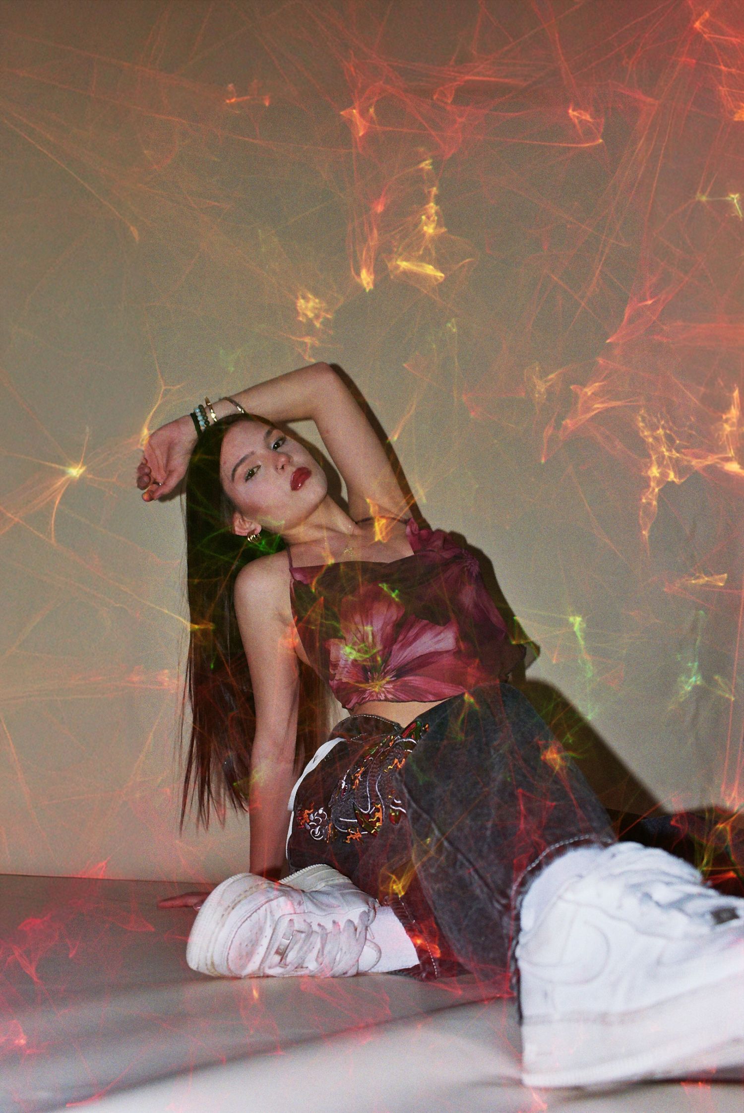 Young woman in colorful mesh top and white sneakers poses on floor against wall with vibrant laser light display.