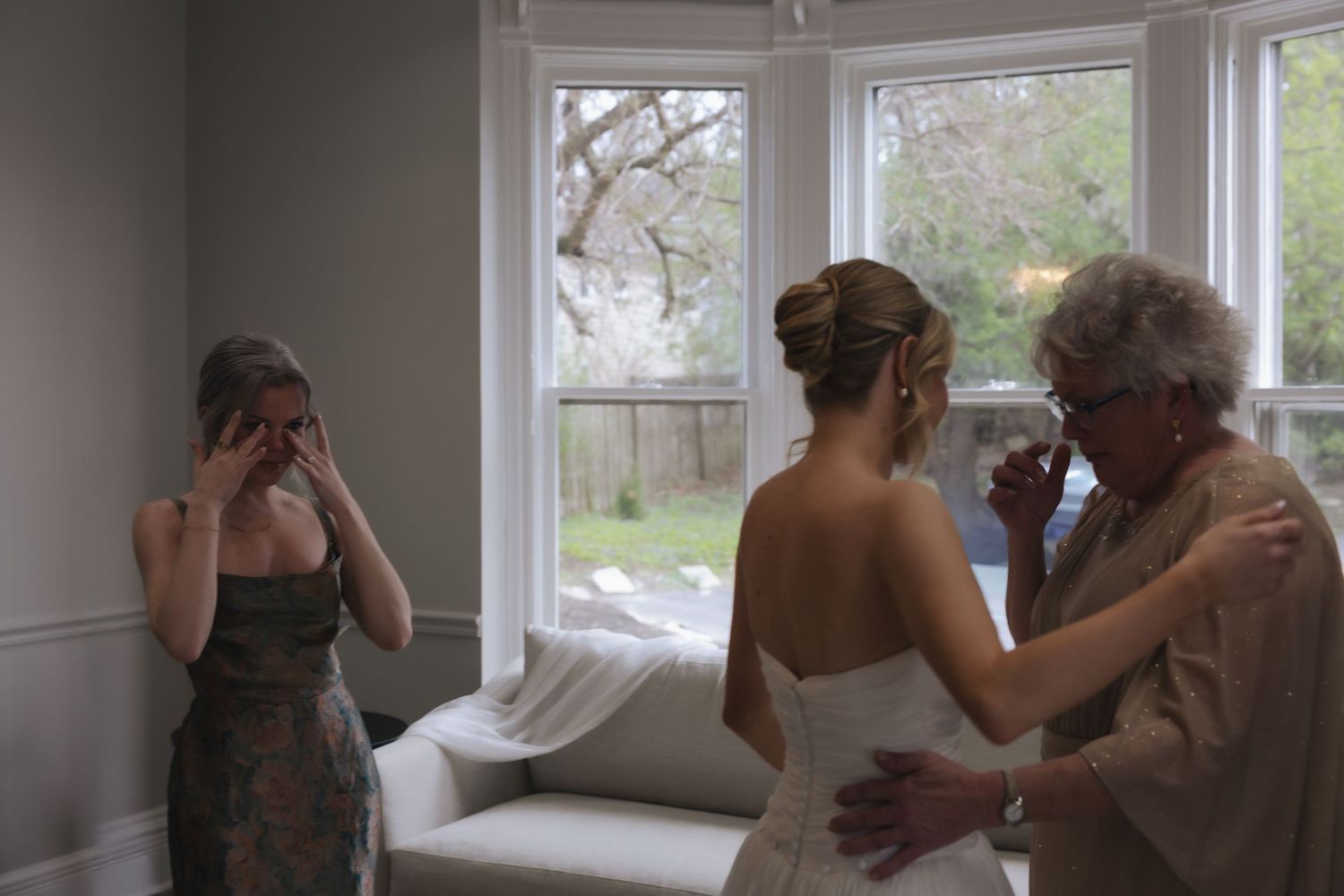 Emotional wedding scene with bride in white gown, tearful bridesmaid, and elderly woman in a bright windowed room.