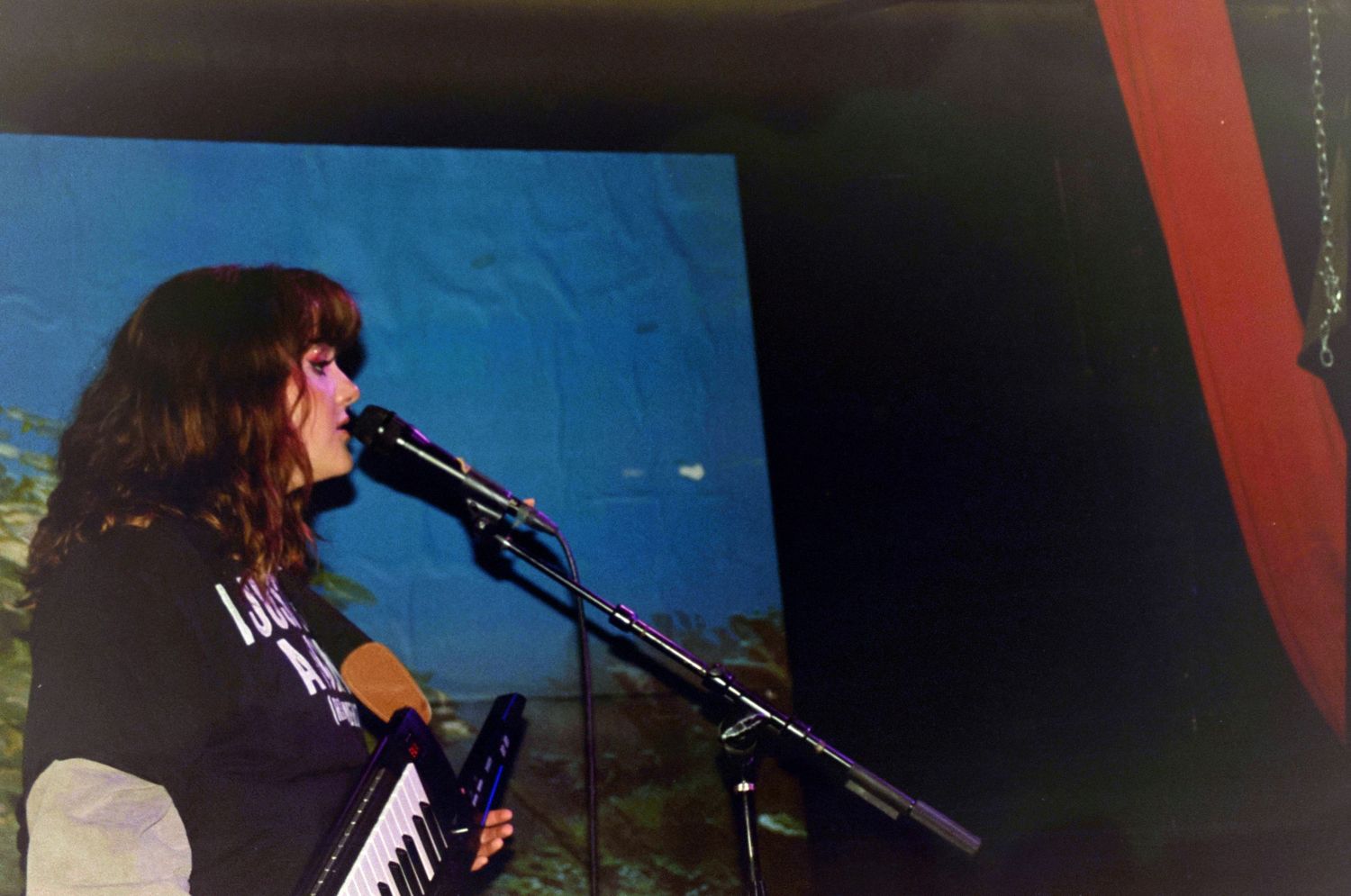Female musician playing keyboard synthesizer at microphone stand in front of blue projected backdrop on stage.