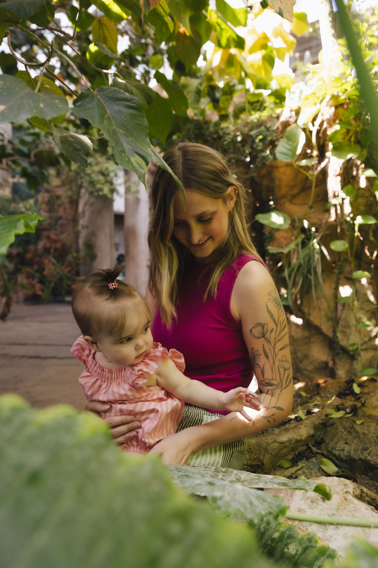 A tattooed young mother smiles at her baby girl in a pink dress, sitting among lush green tropical plants.