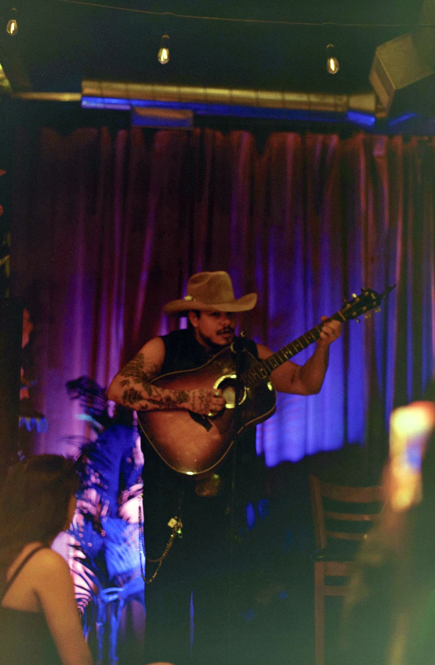 Tattooed male musician performing on stage playing acoustic guitar wearing cowboy hat in dimly lit venue.