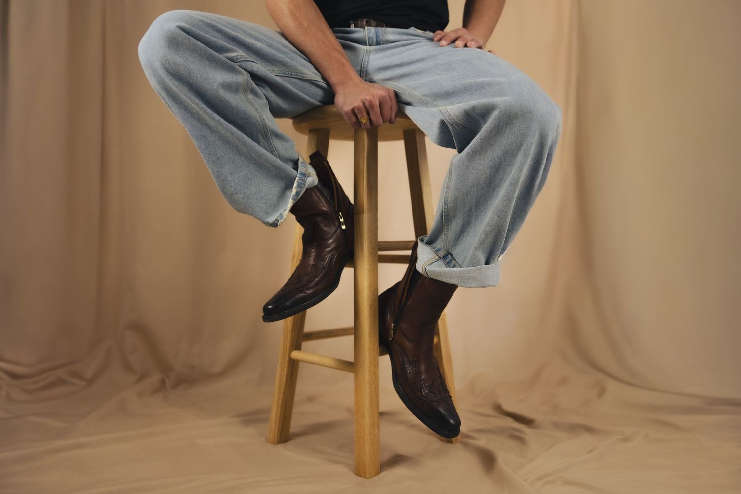 Person sitting on wooden stool wearing light wash jeans and dark brown leather cowboy boots against beige backdrop.