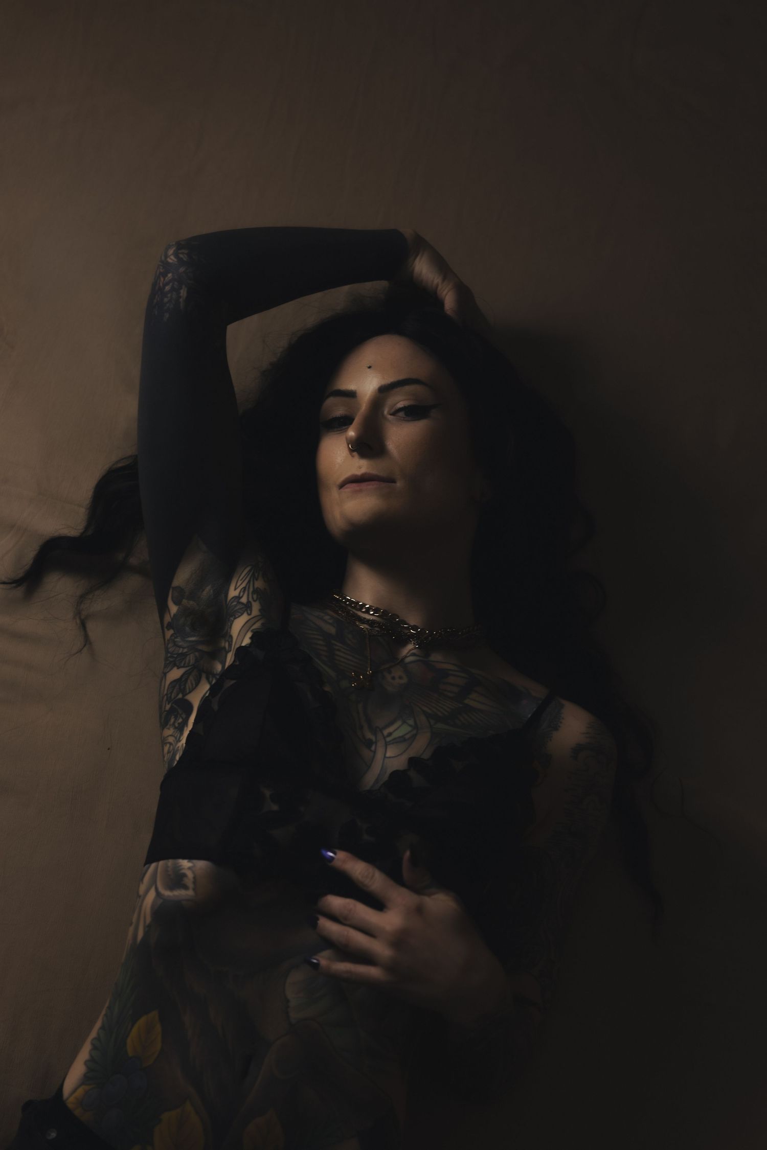 A tattooed woman with dark hair poses dramatically against a dark wall, wearing an ornate patterned garment.