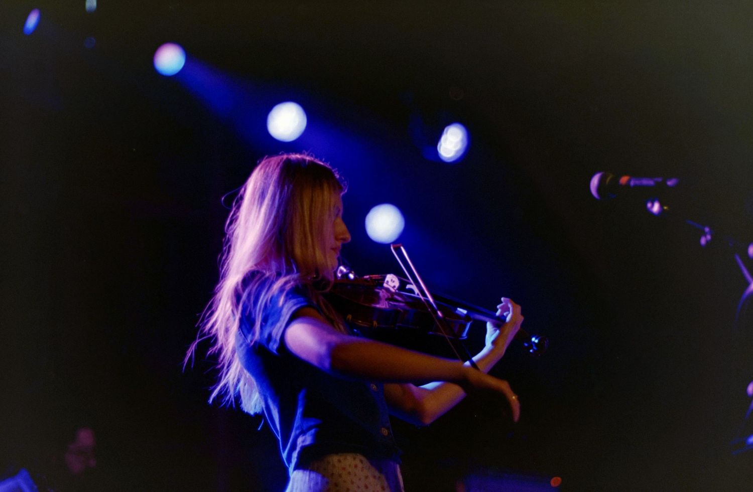 A female violinist performs on stage under blue and purple spotlights during a live concert.