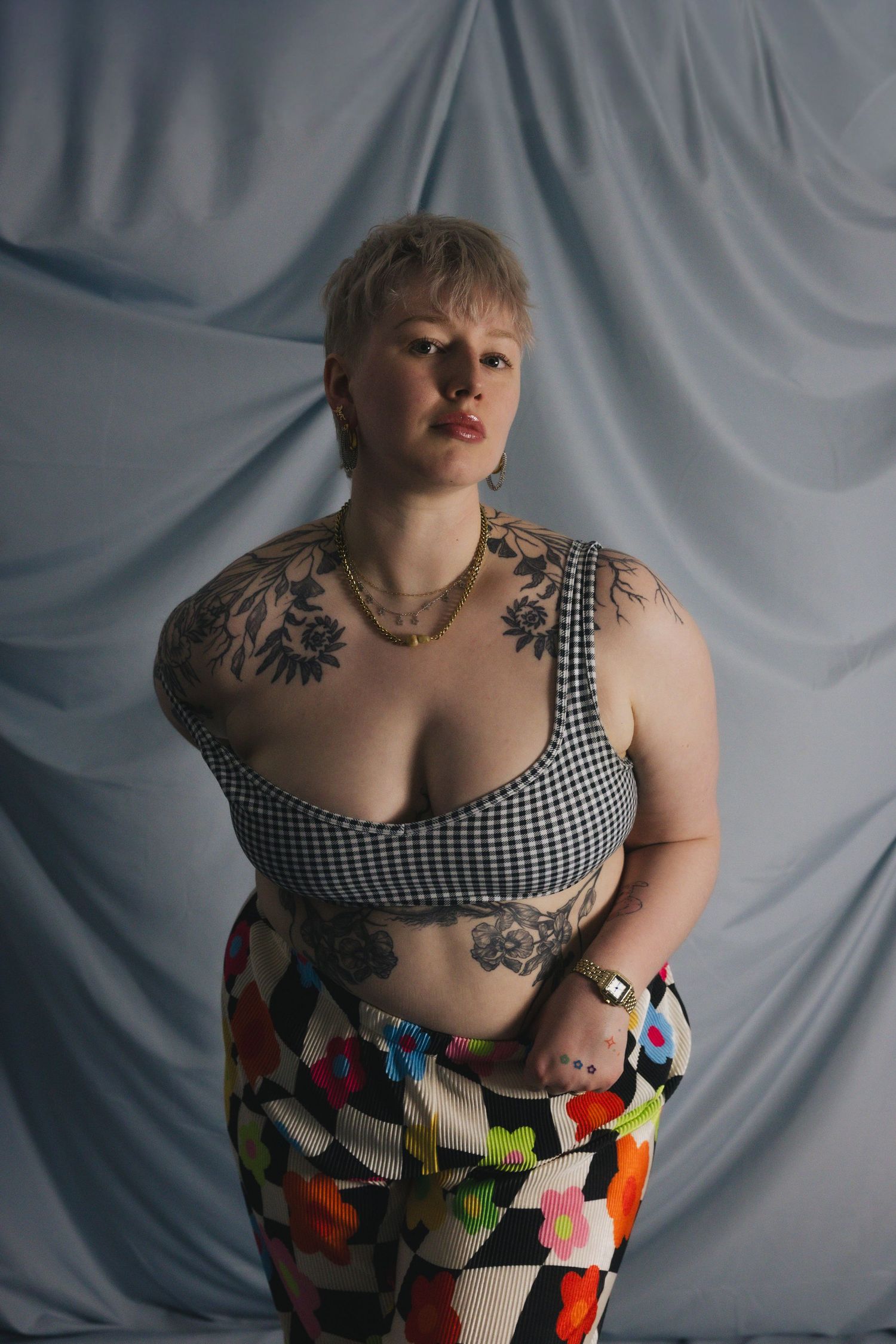 Plus-size woman with tattoos wearing a checkered crop top and colorful floral pants posing against a draped gray backdrop.