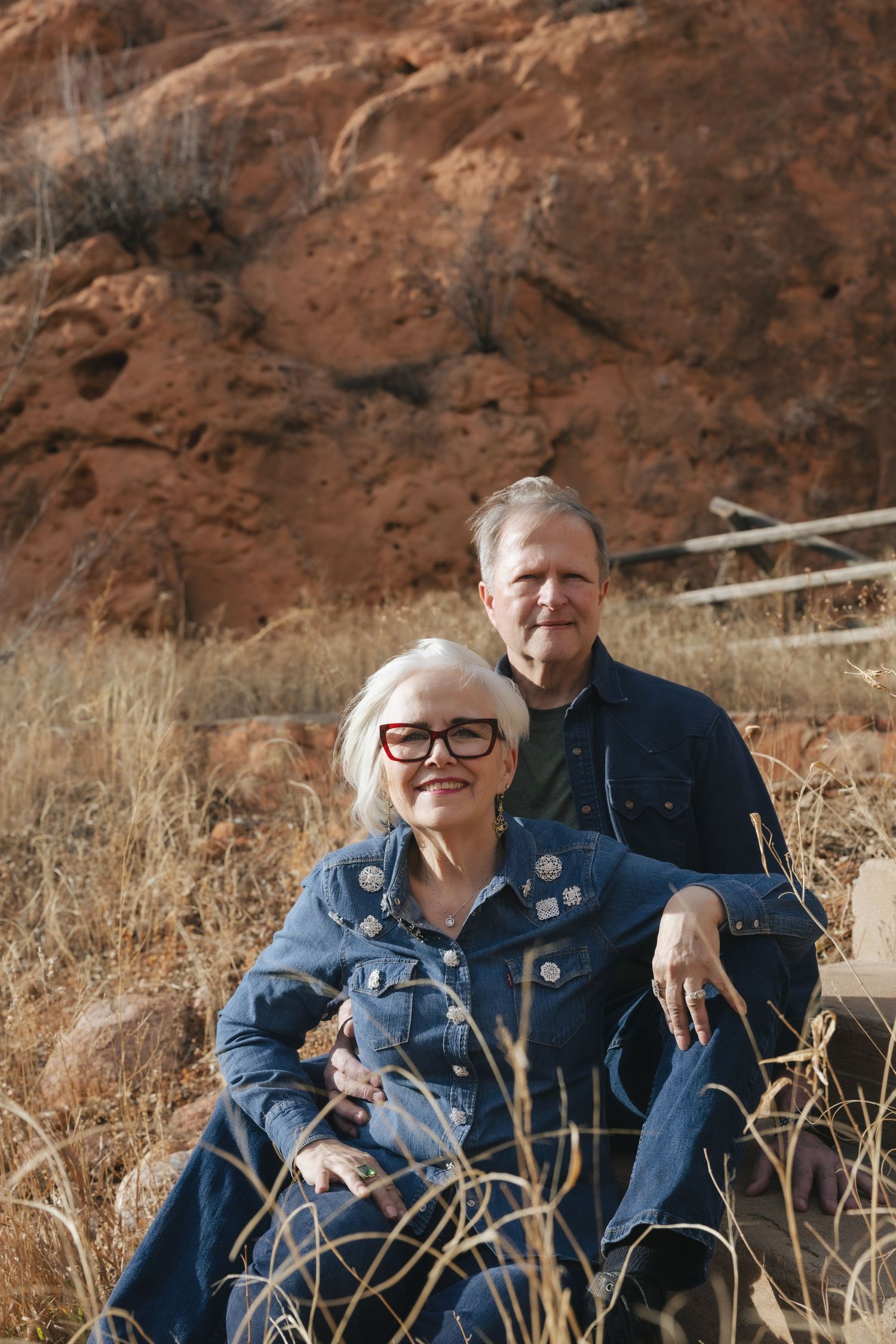 Older couple in denim sitting outdoors near red rock formations, smiling warmly in natural desert landscape.