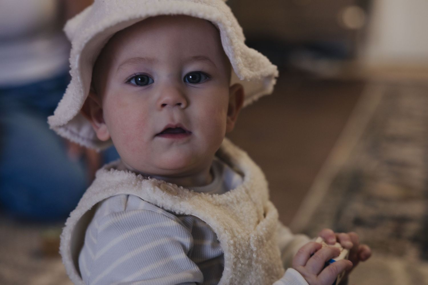 Adorable baby wearing a white fuzzy hat and vest, looking curiously at the camera with big brown eyes.