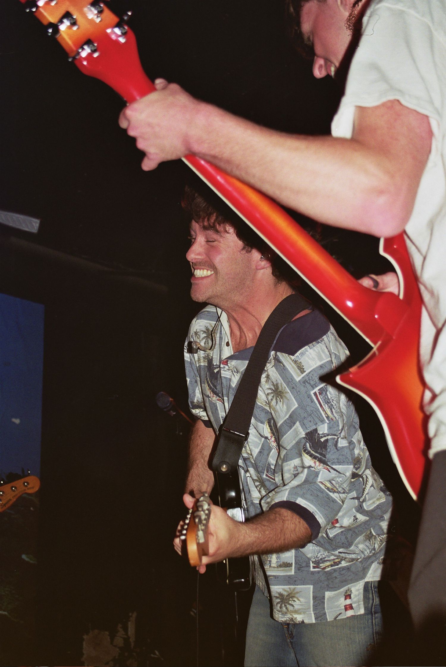 Two musicians performing live on stage, one smiling in a Hawaiian shirt playing guitar while another holds a red electric guitar overhead.