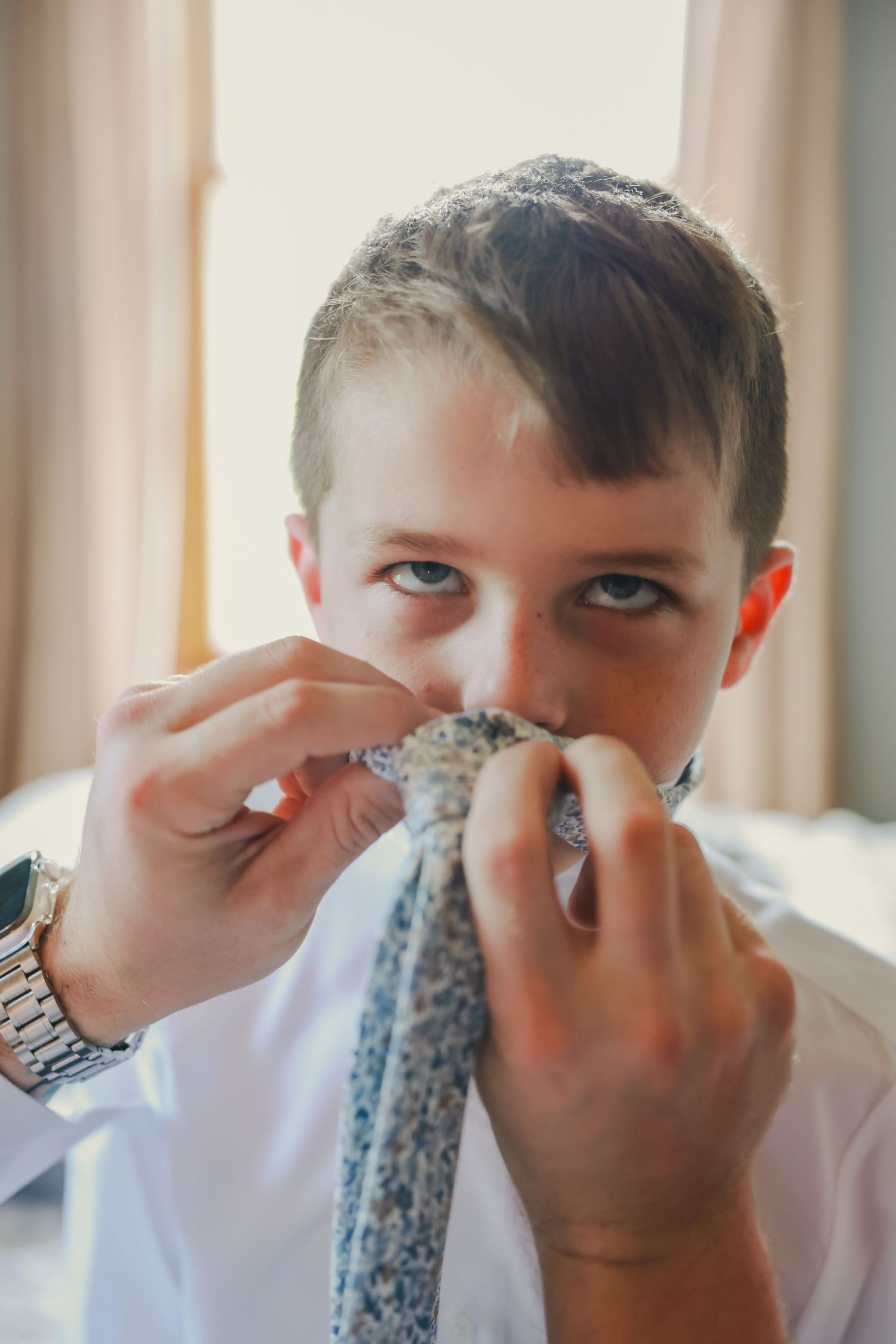 Young person in white shirt playfully holding and smelling a patterned blue necktie by a window.