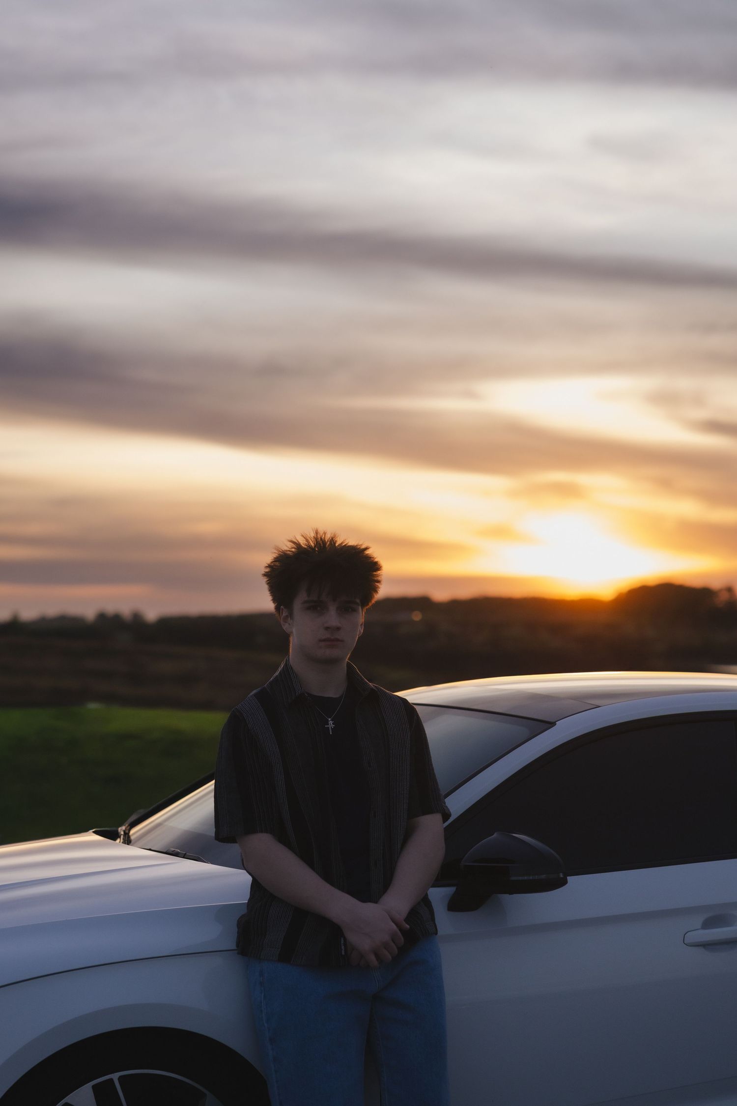 Young man leaning against a white car during a stunning golden sunset over green fields.