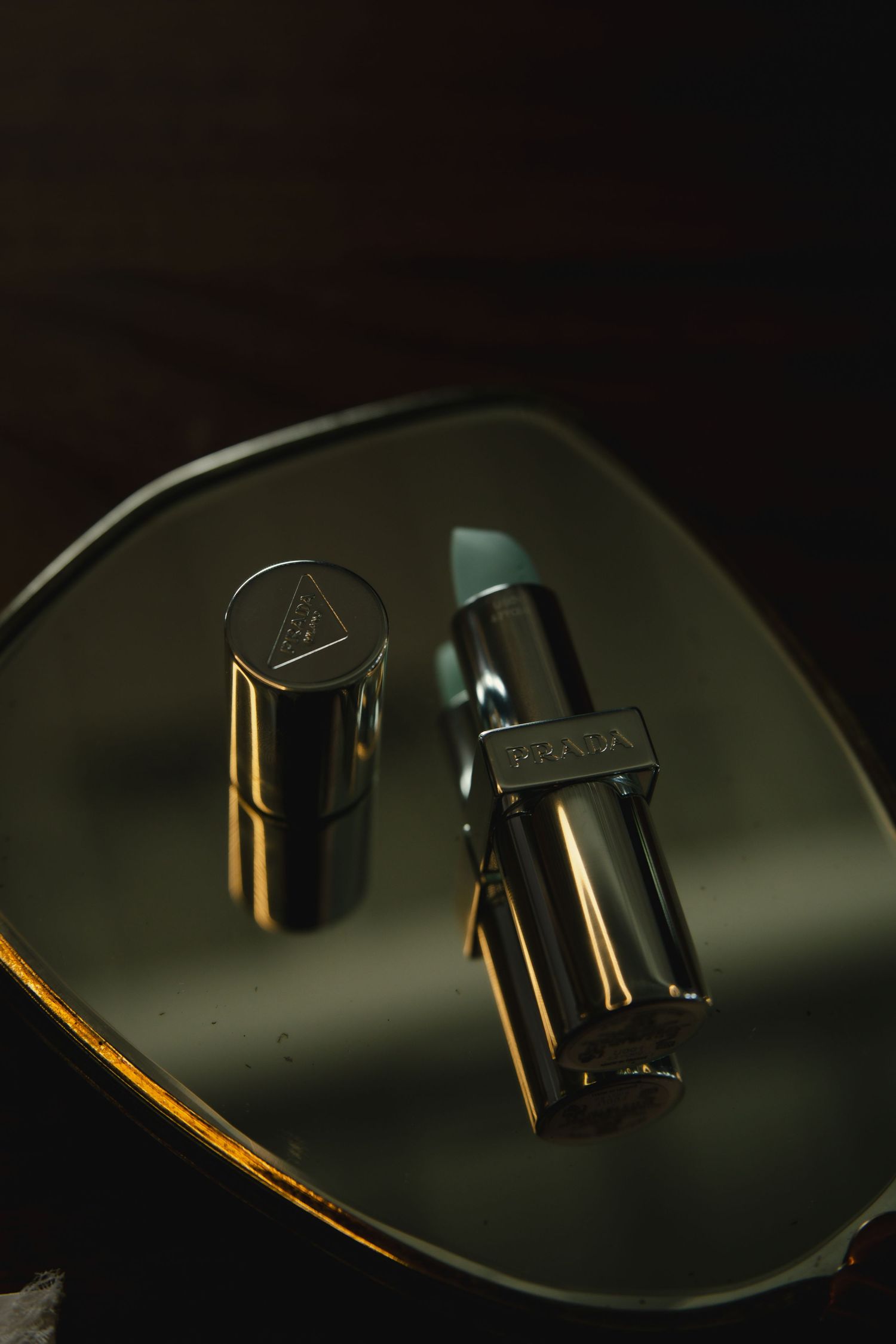Two black luxury lipstick tubes with metallic gold accents reflecting on a dark glossy surface in moody lighting.
