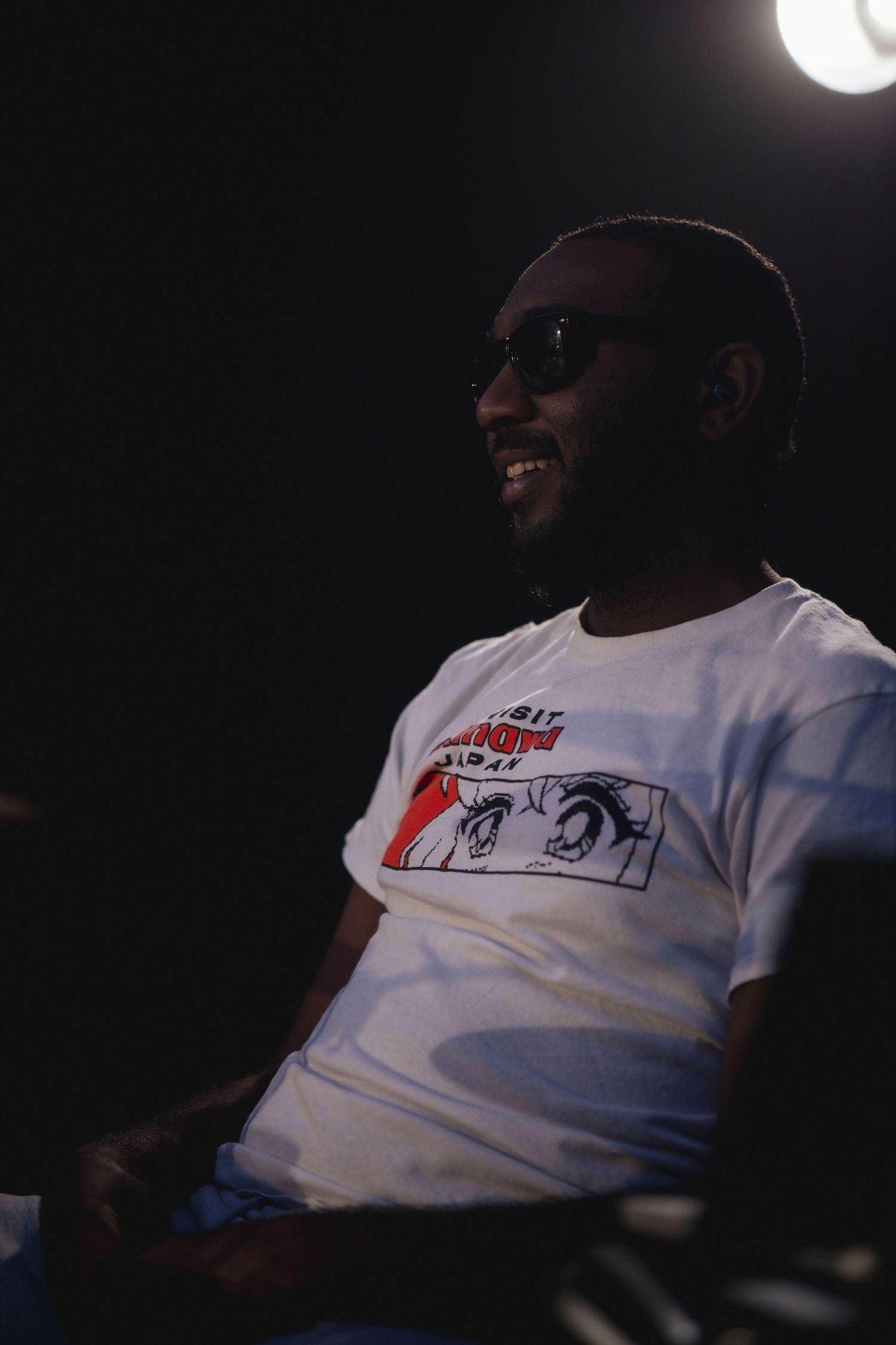 A person in sunglasses and white graphic t-shirt smiles while sitting in a dark setting.