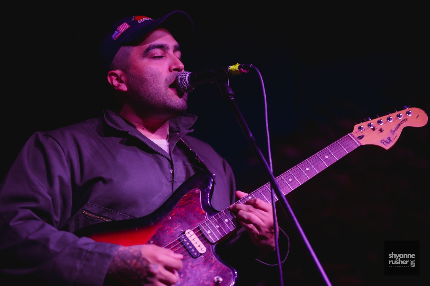 A musician performs on stage with an electric guitar under dramatic red lighting while singing into a microphone.