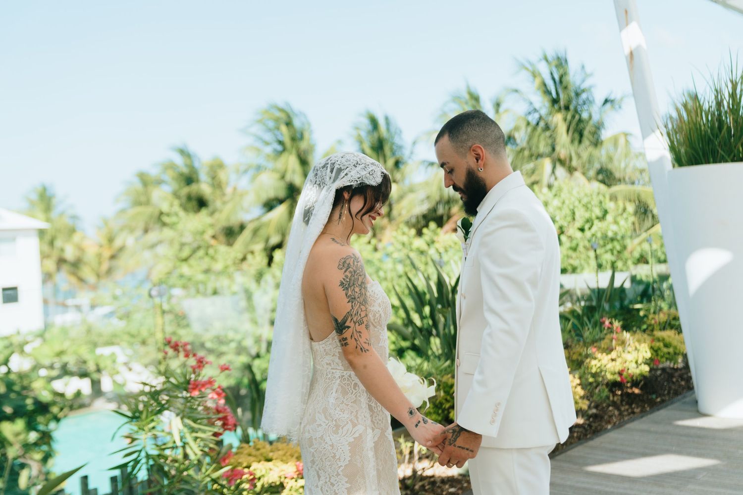 Bride and groom holding hands in a tropical garden, bride wearing lace gown with veil, groom in white suit.