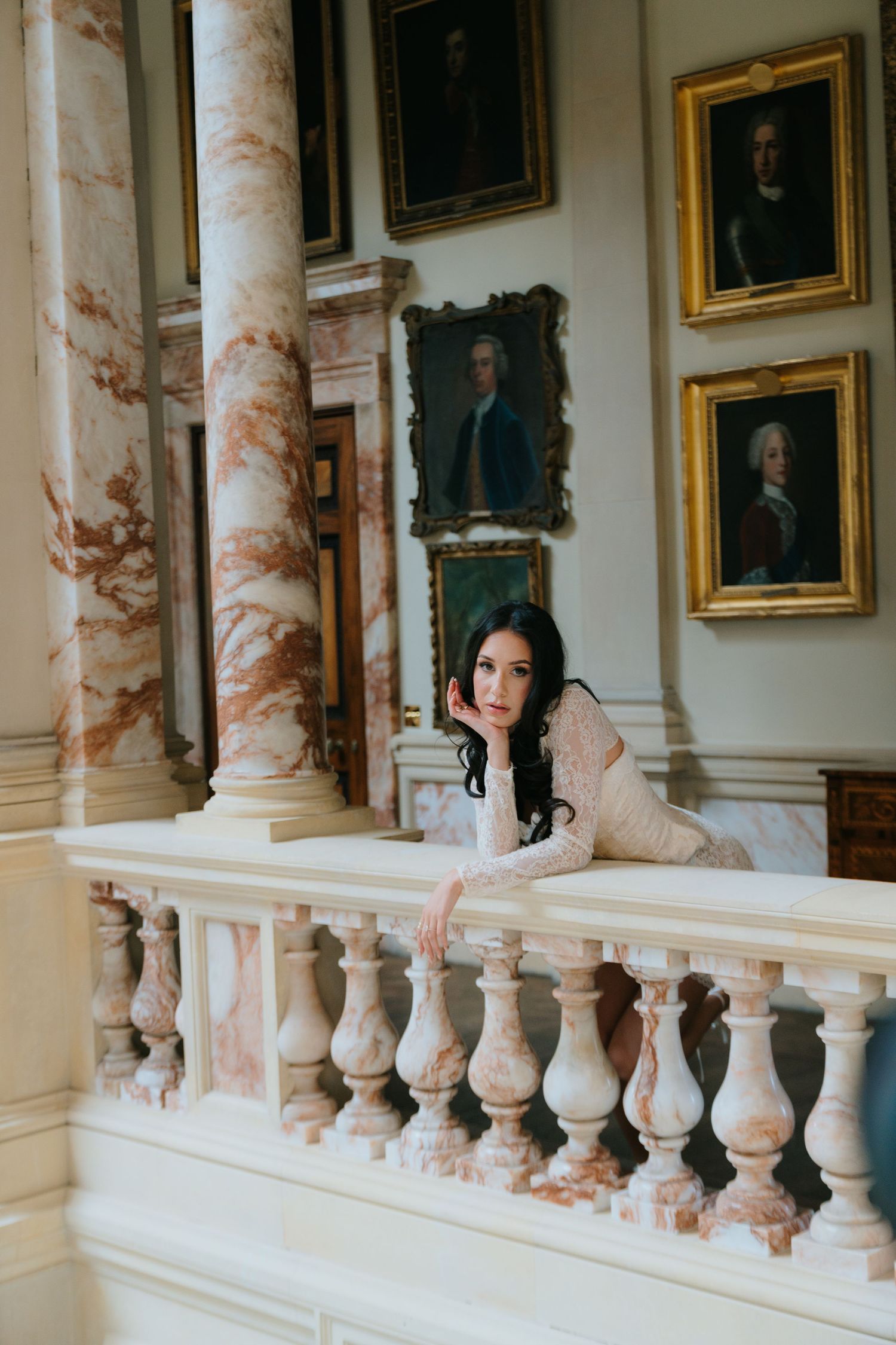 Elegant brunette woman leans on marble balustrade surrounded by classical columns and portrait paintings.