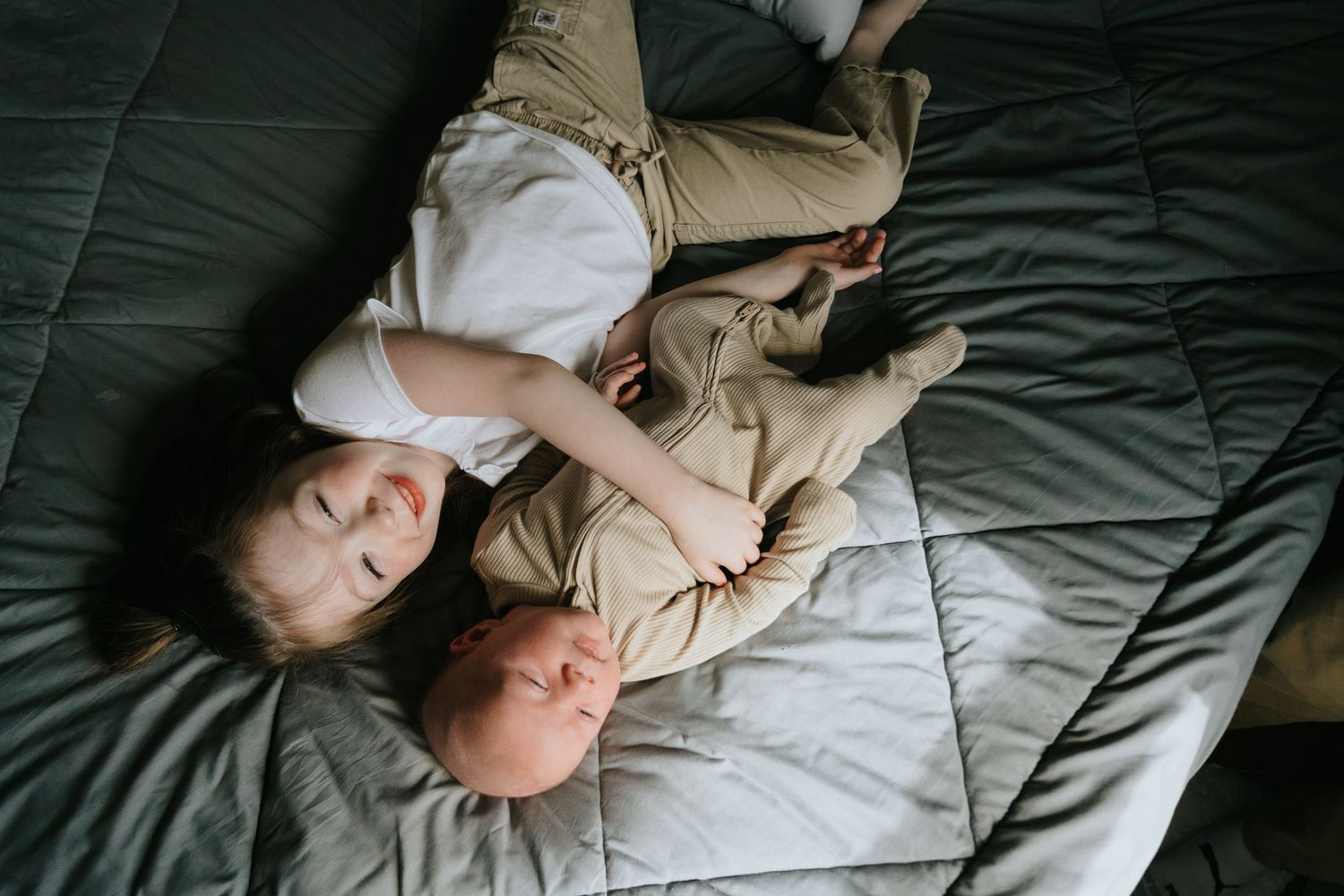 Toddler and newborn baby lying together on a gray bed, older sibling gently holding infant.