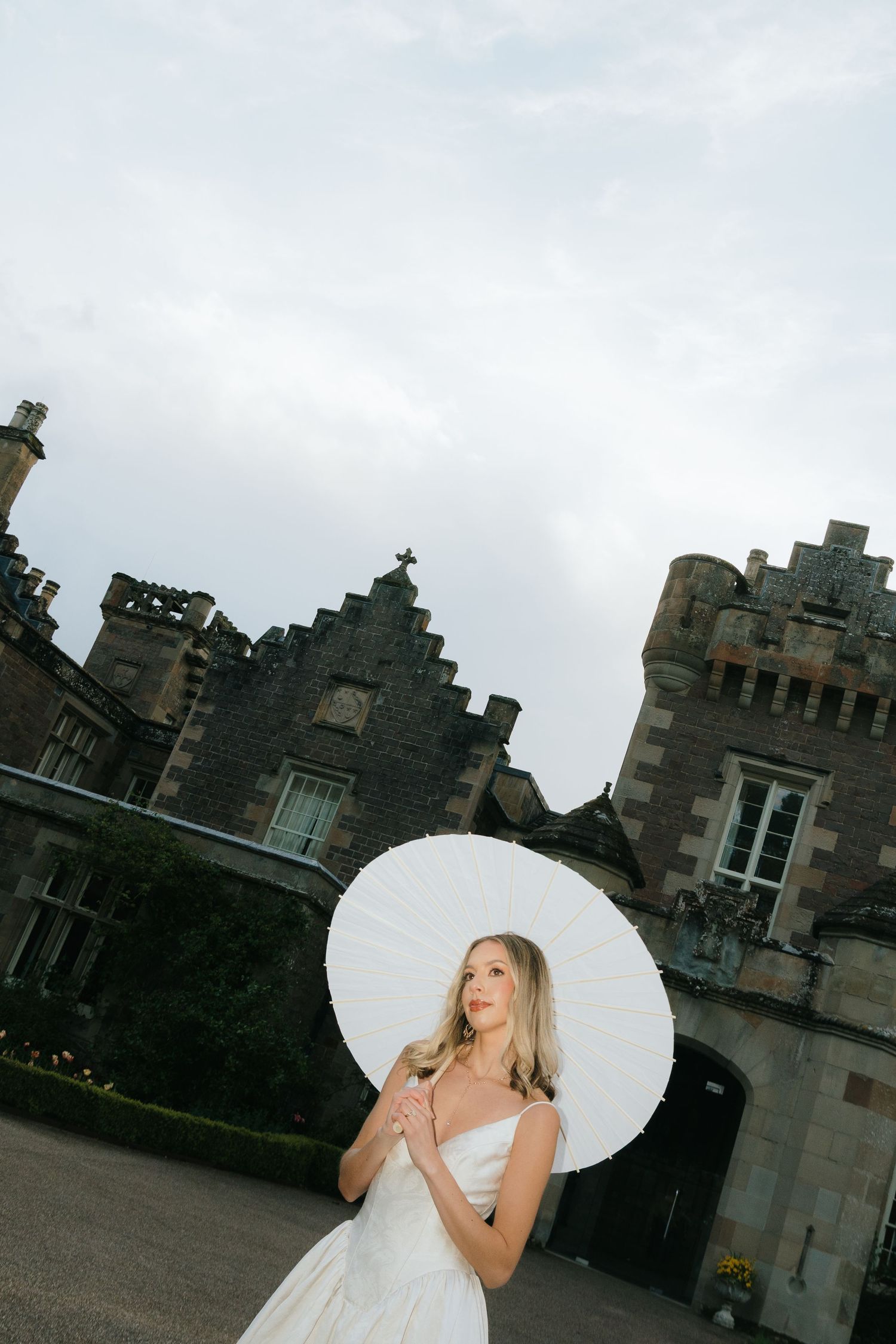Bride in white dress holding white parasol umbrella standing before a historic stone castle under cloudy skies.