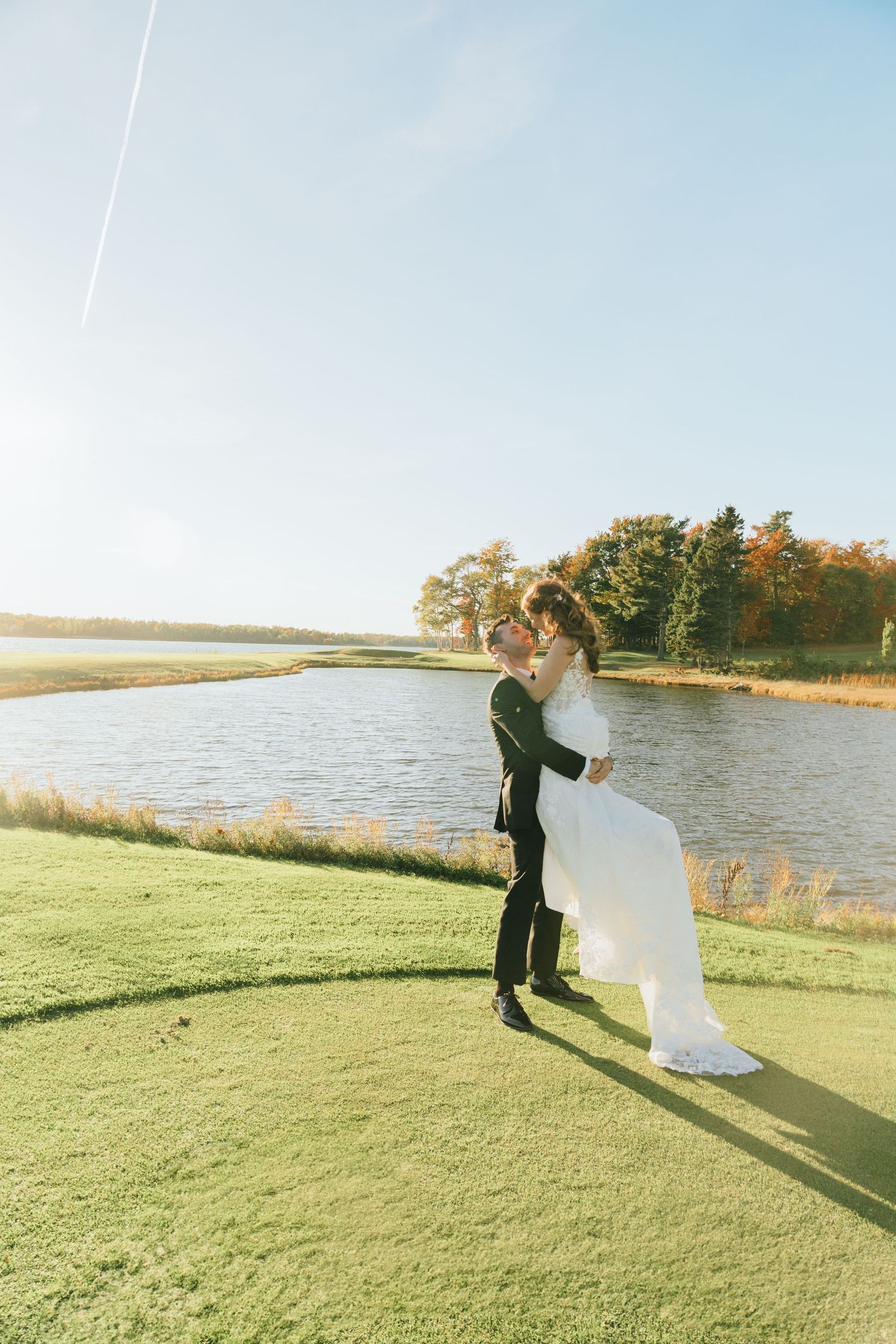 Groom lifting bride in white gown by a sunlit lake on green grass, autumn trees in background.