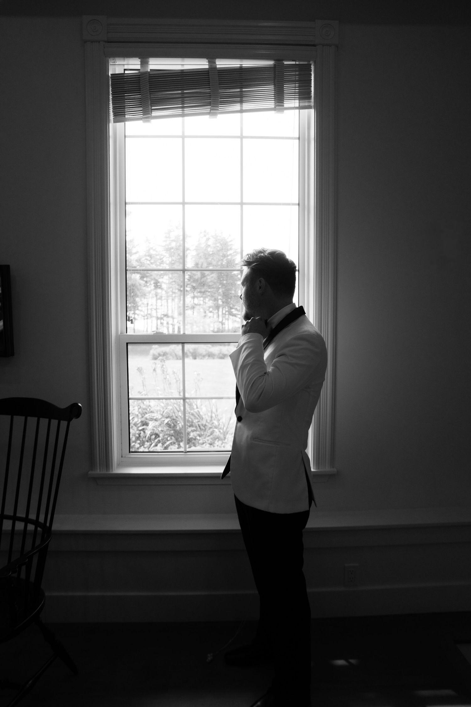 Black and white photo of a groom in a white jacket adjusting his tie while gazing out a large window.