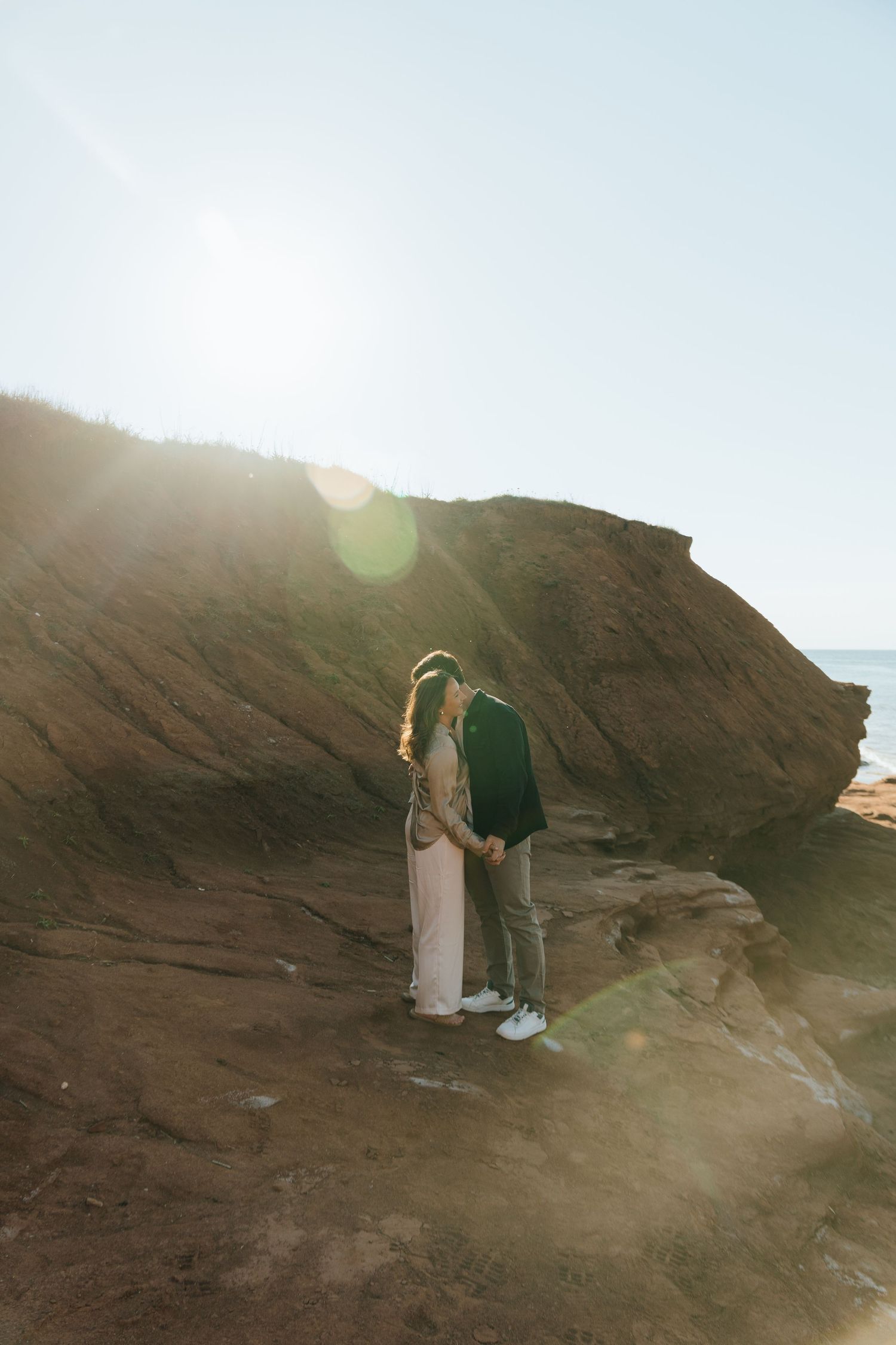 Couple sharing a romantic moment on rocky coastal cliffs with golden sunlight and ocean backdrop.