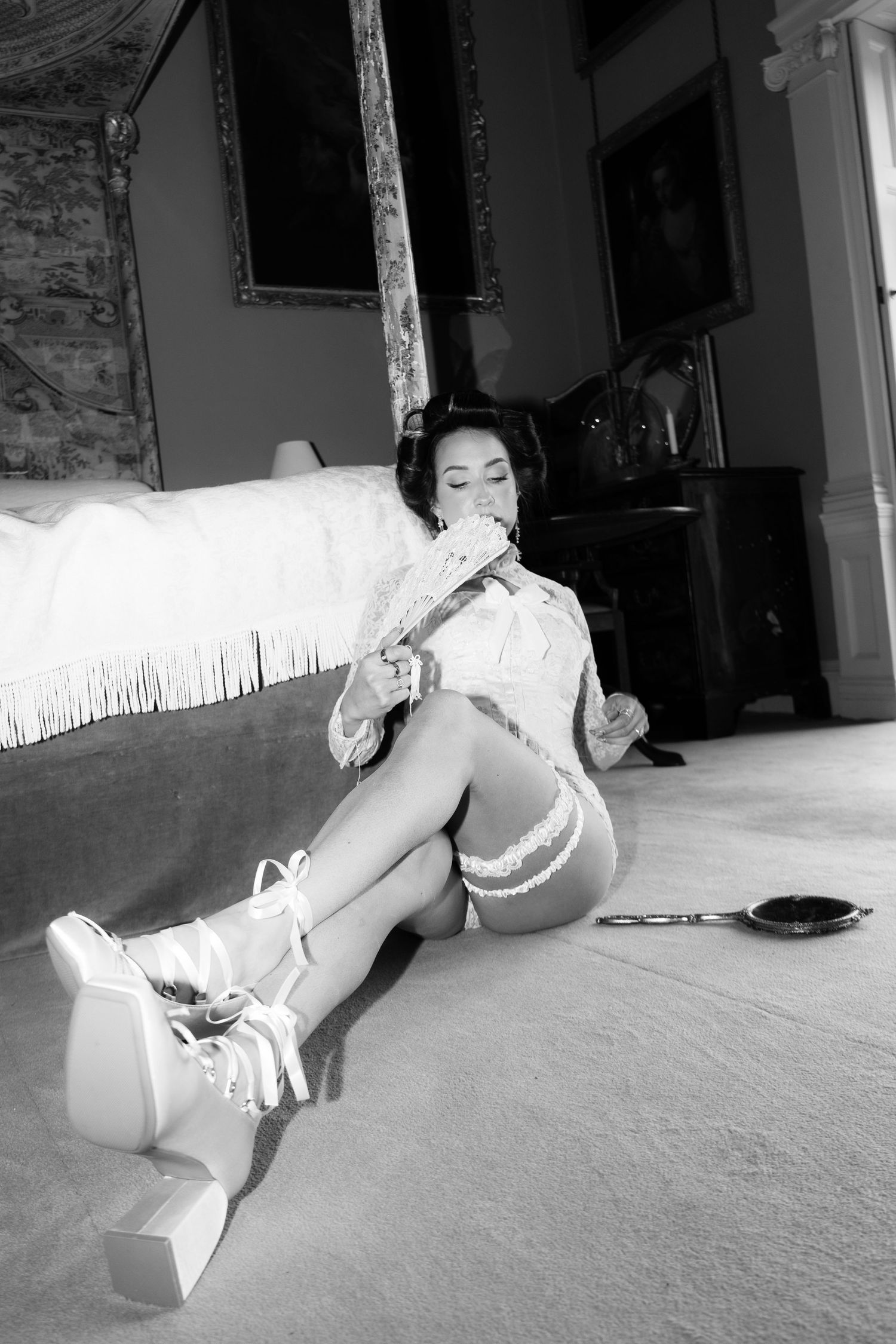 Black and white photo of a woman in shorts and platform shoes sitting on floor leaning against a bed holding a fan.