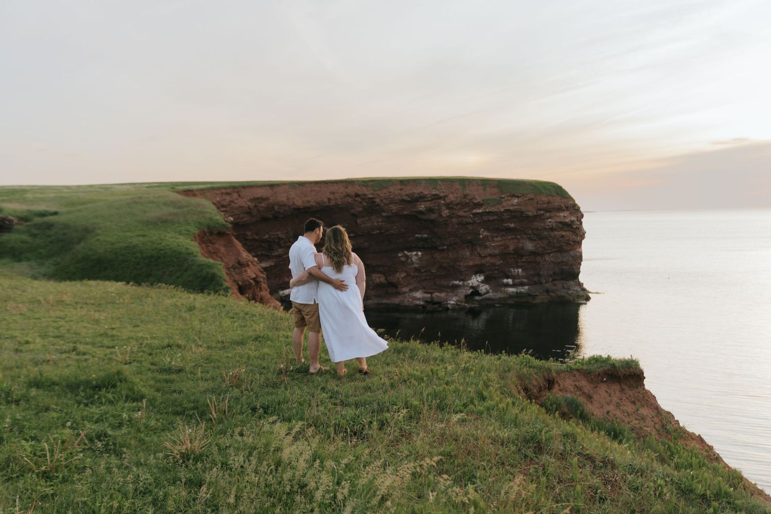 Couple in white outfits sitting together on a scenic coastal cliff overlooking the ocean at sunset.
