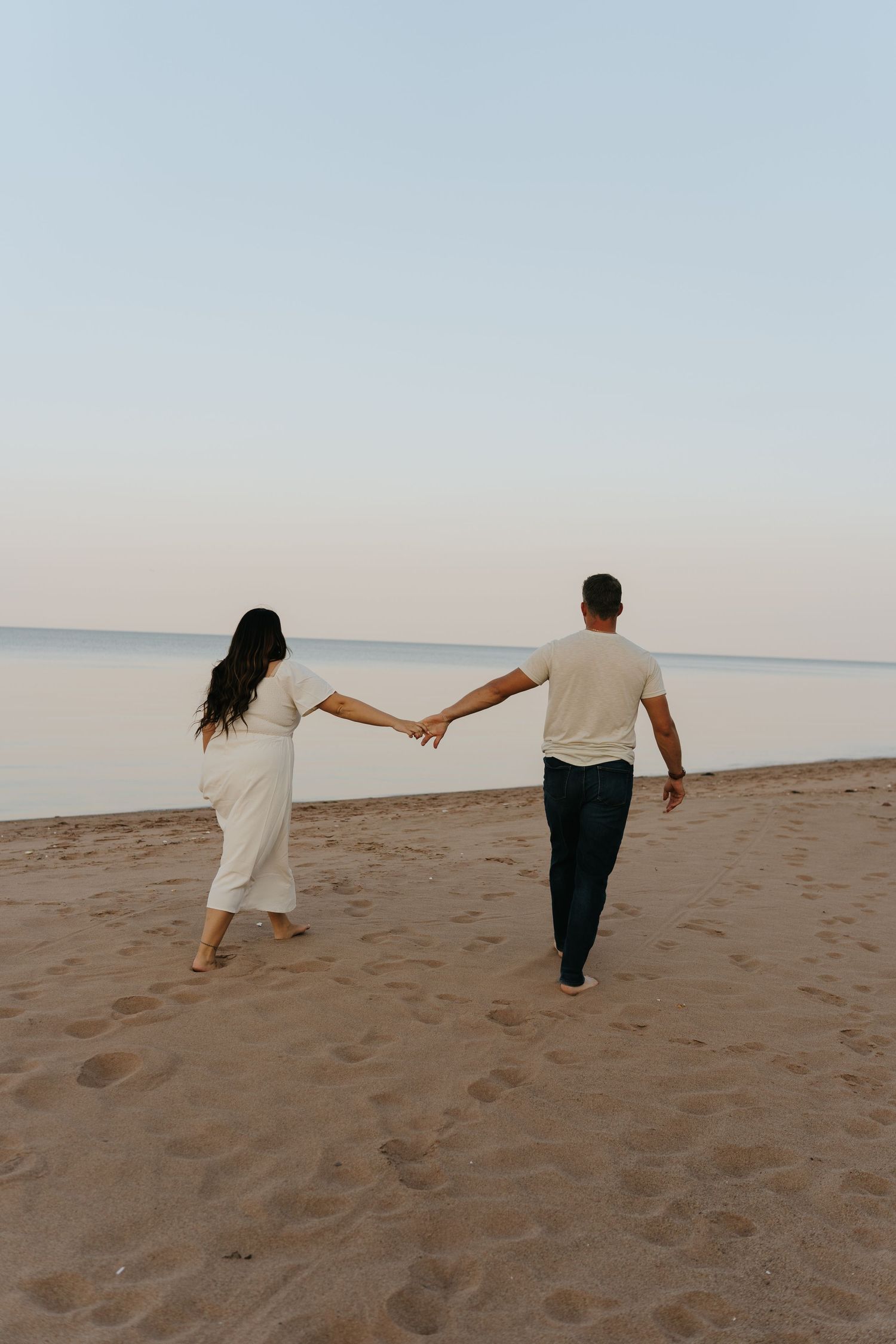 Two people hold hands while walking along a sandy beach at dusk.