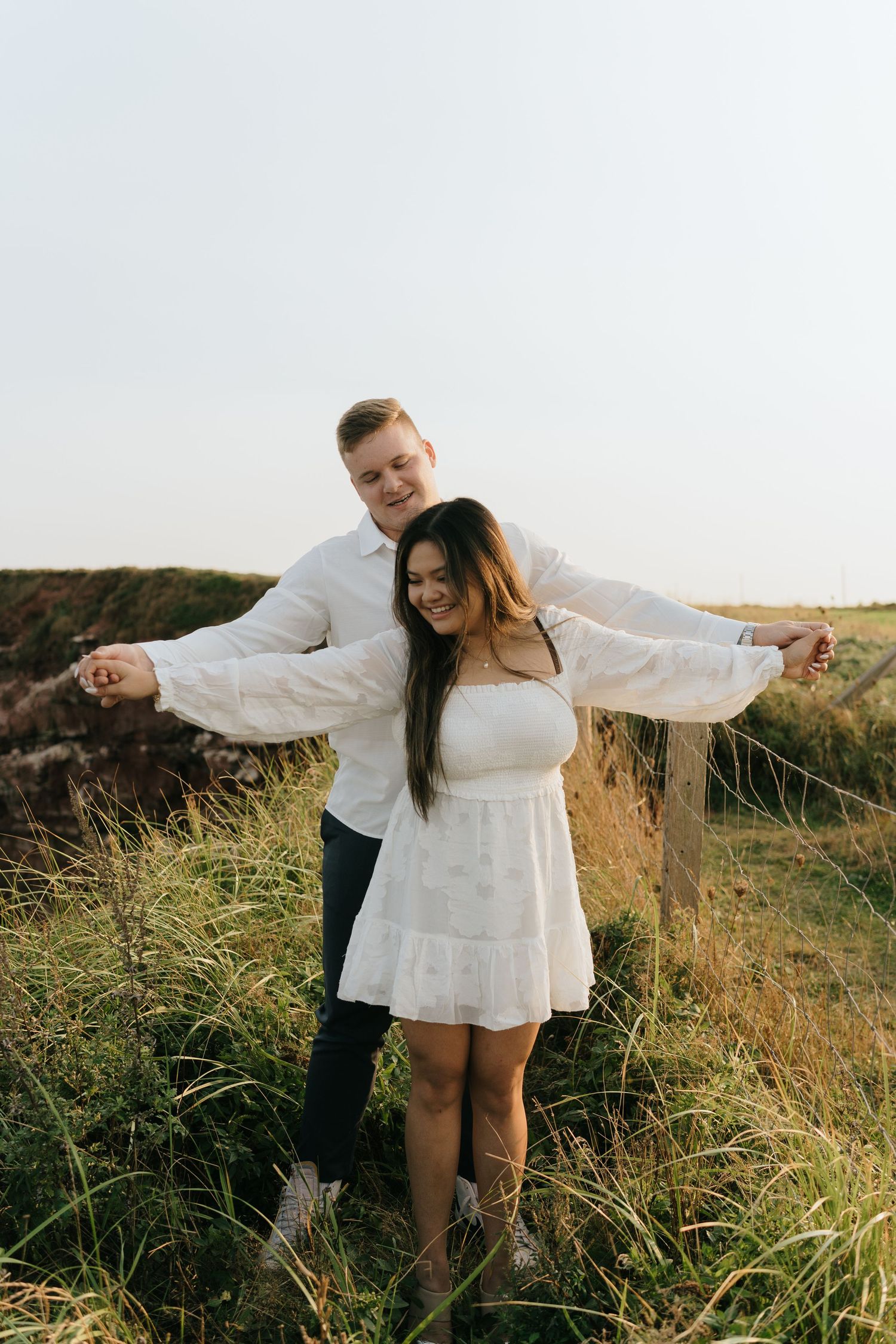 Couple in white clothing posing playfully on a grassy hillside at sunset.