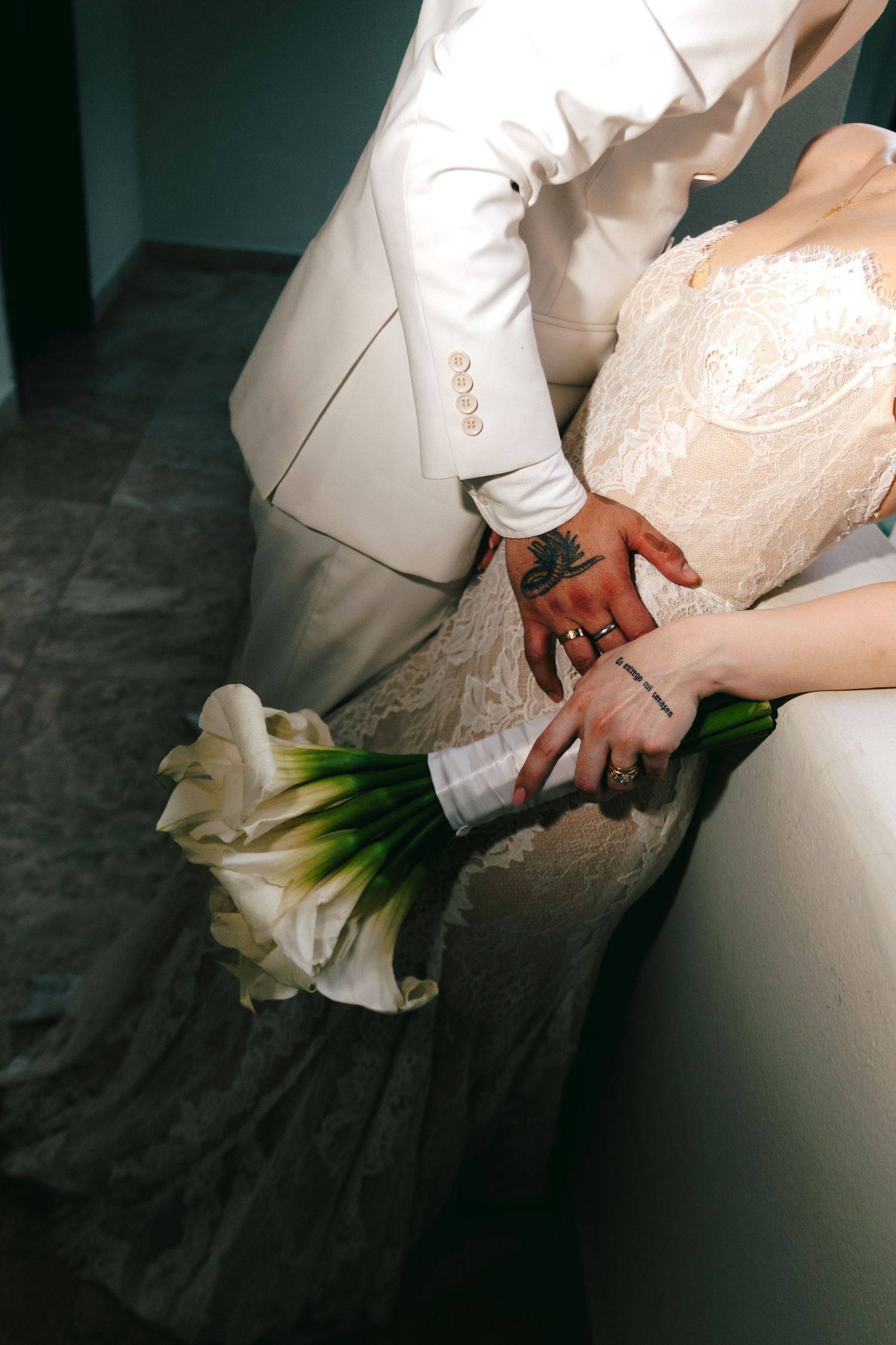 Bride and groom holding white calla lily bouquet, groom with tattooed hand wearing white suit, bride in lace wedding dress.
