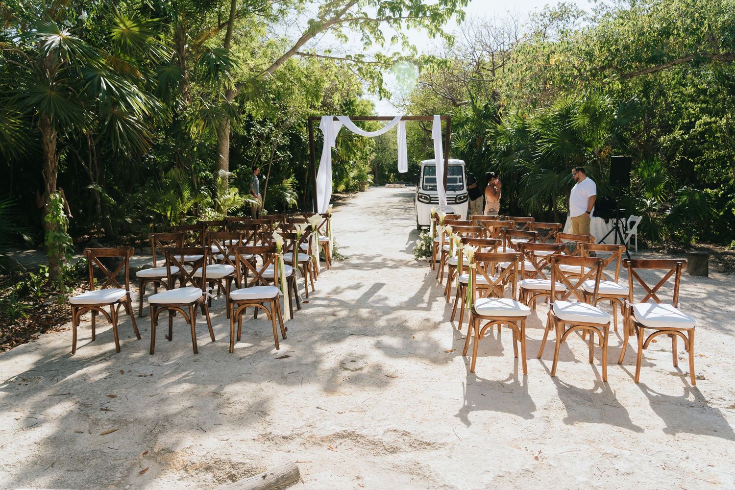 Outdoor beach wedding ceremony setup with wooden cross-back chairs, white floral arch, and lush tropical greenery.