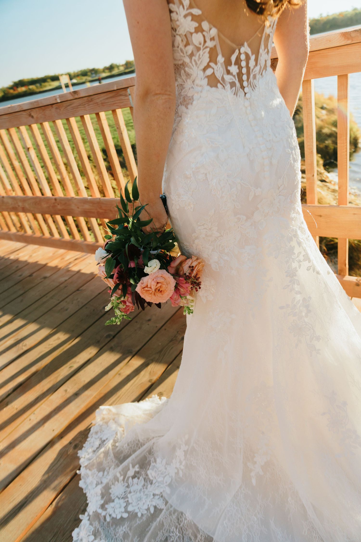 Bride in a lace mermaid wedding gown holding a pink floral bouquet on a wooden deck outdoors.