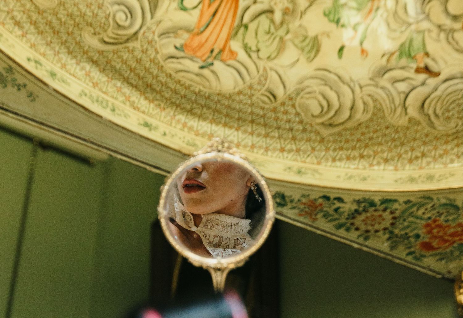 A reflection of a woman in a decorative handheld mirror against an ornate painted ceiling.