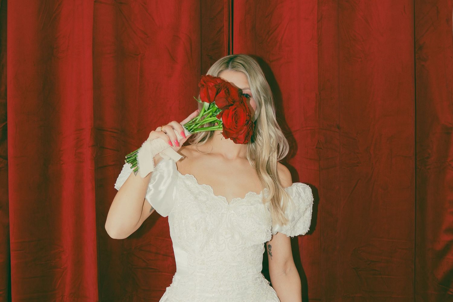 Romantic bridal portrait with red roses against crimson curtain backdrop.
