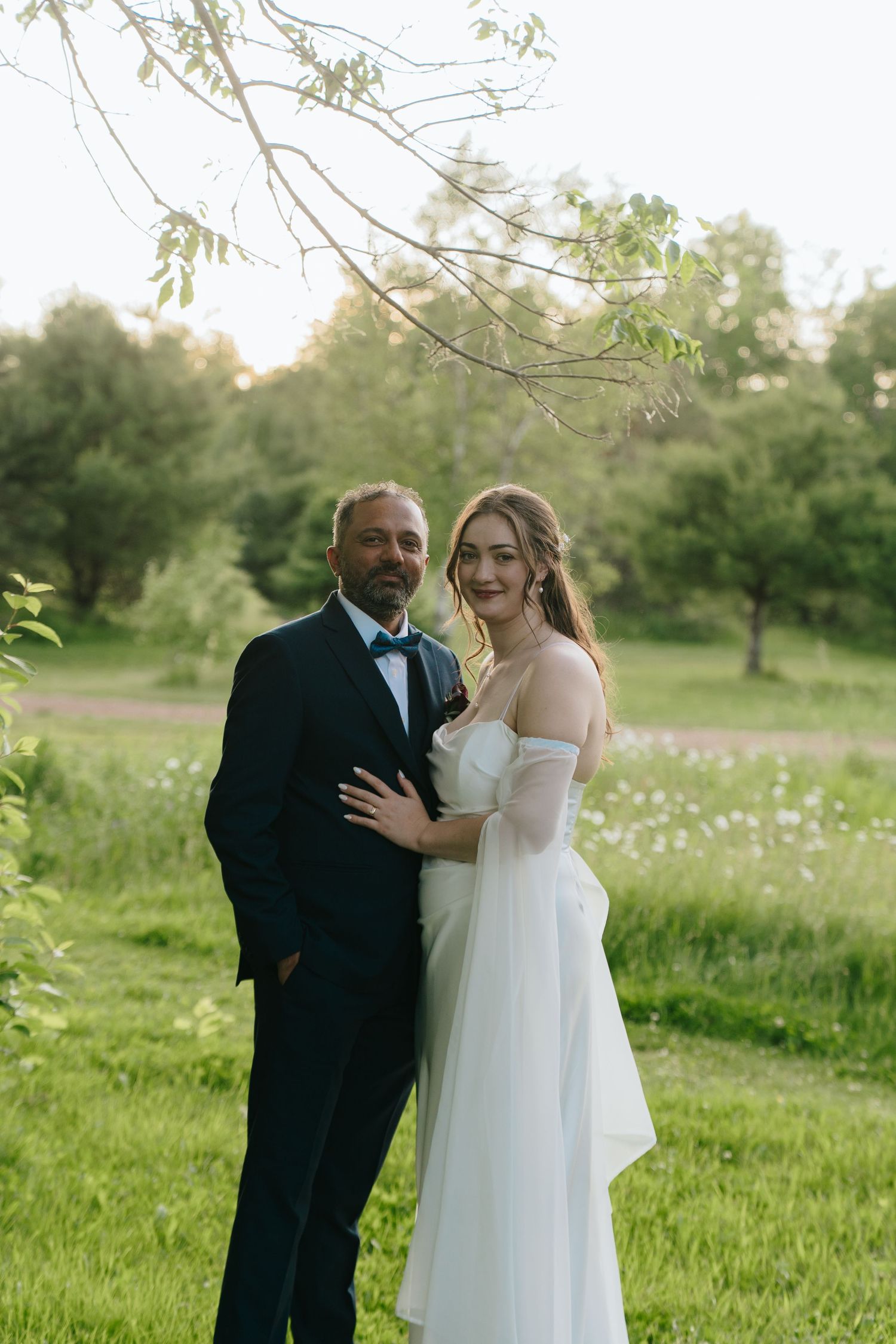 A couple poses together in a spring meadow with blooming wildflowers and trees.