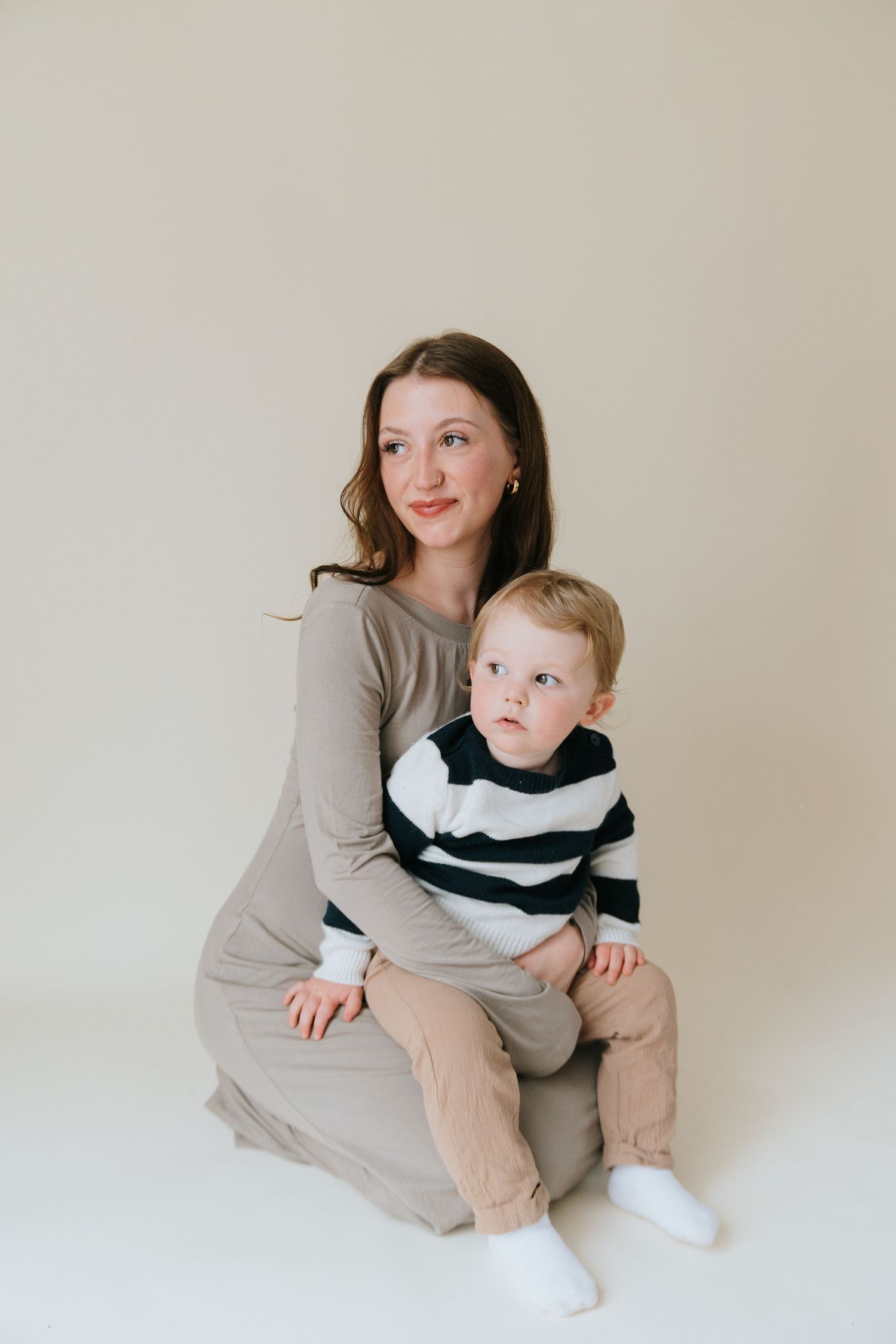 A young mother in a taupe dress sits on the floor holding her toddler boy in a striped sweater against a neutral background.