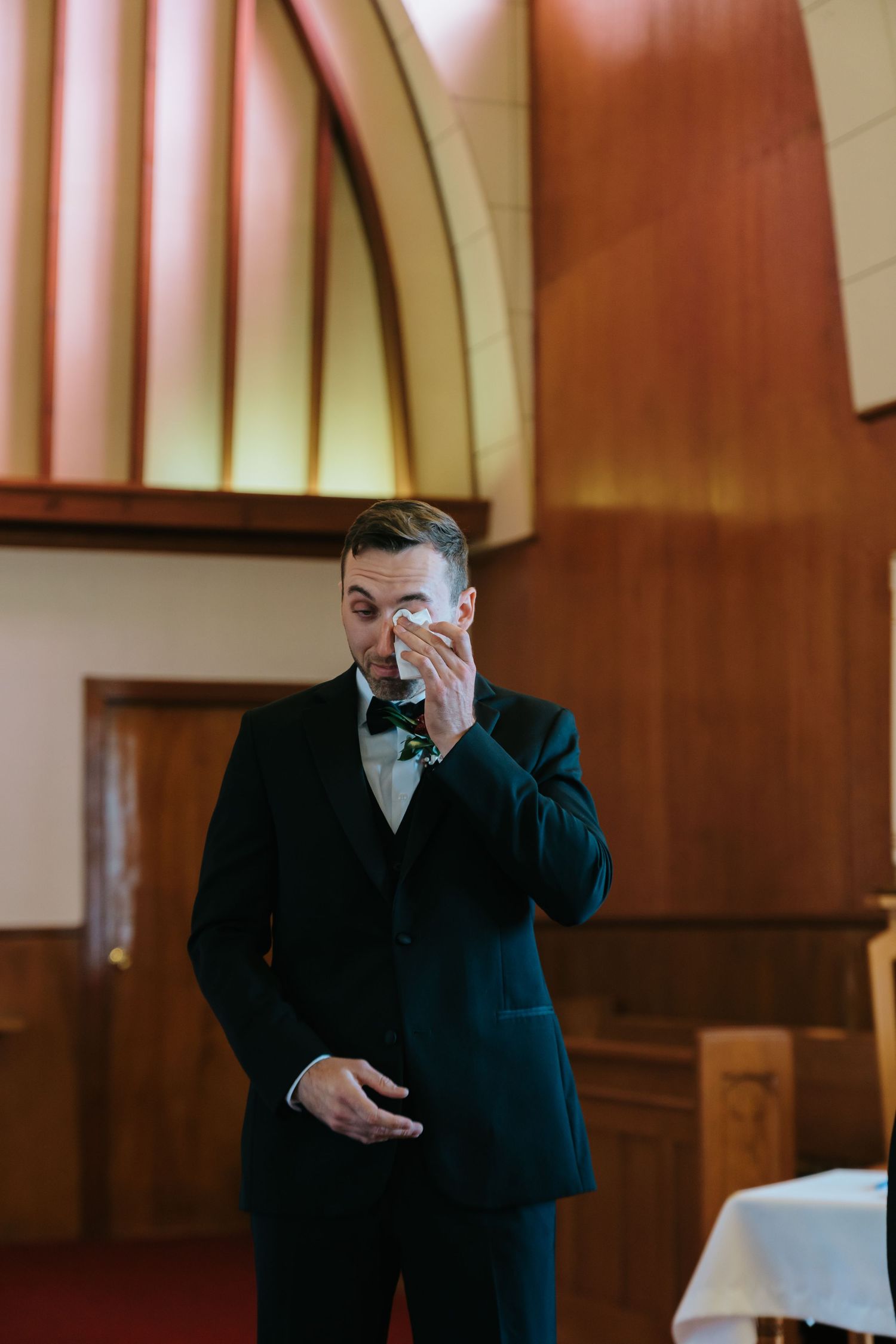 Emotional groom in black tuxedo wiping tears with handkerchief inside a church during wedding ceremony.