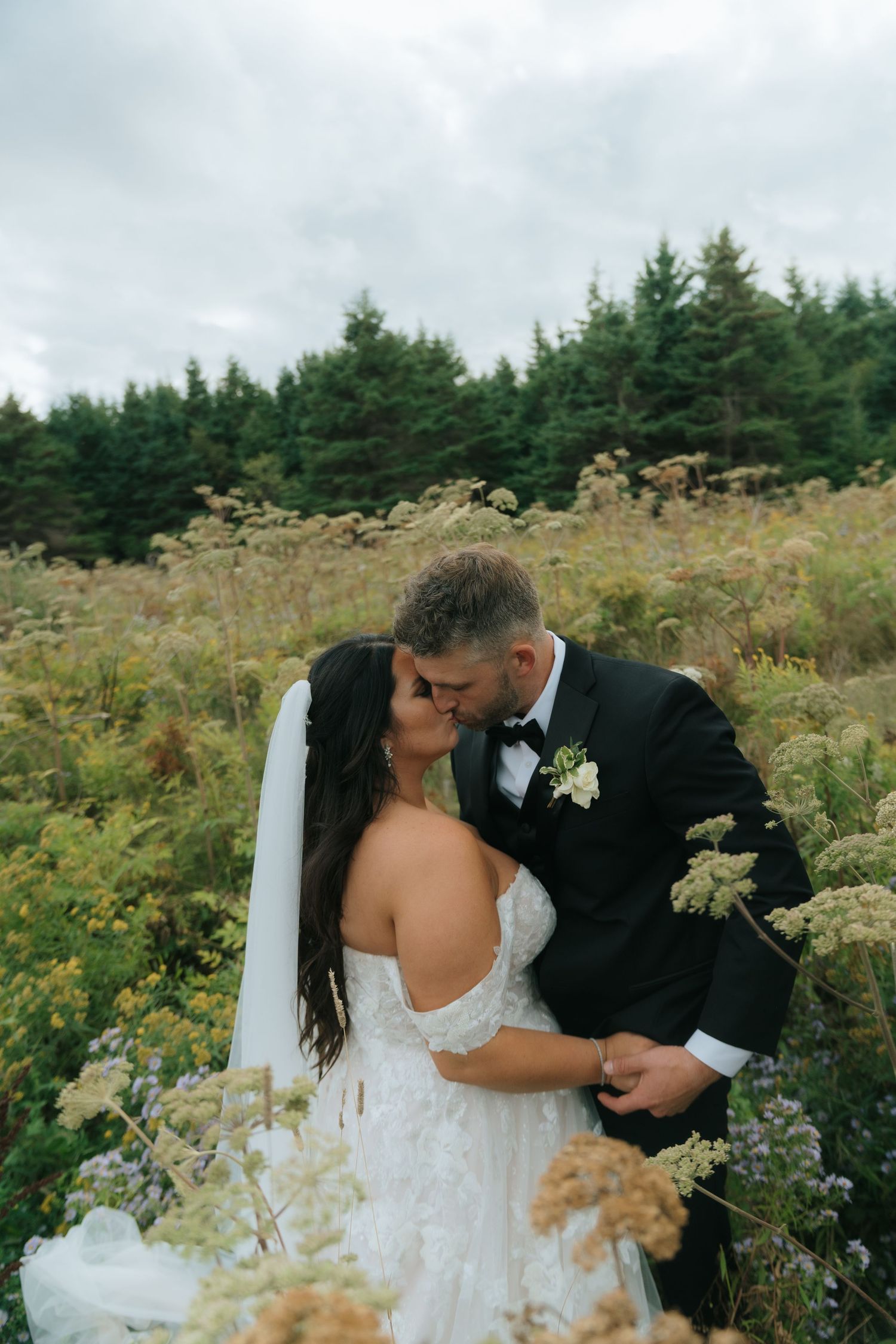 Wedding couple embraces in field of wildflowers with evergreen forest backdrop.